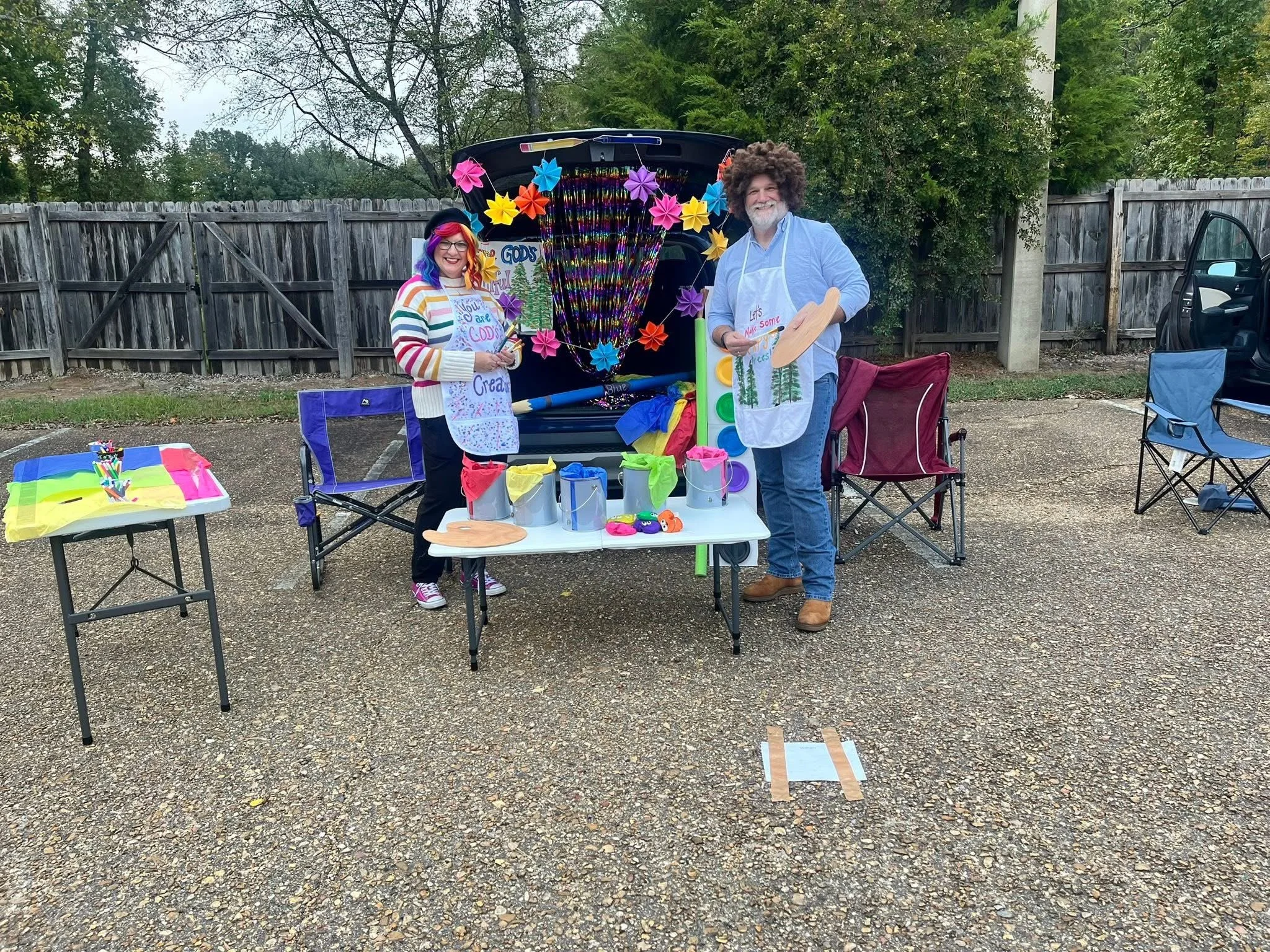 Two people dressed in colorful costumes stand behind a table with craft supplies in an outdoor parking lot, with a truck decorated with paper flowers behind them.