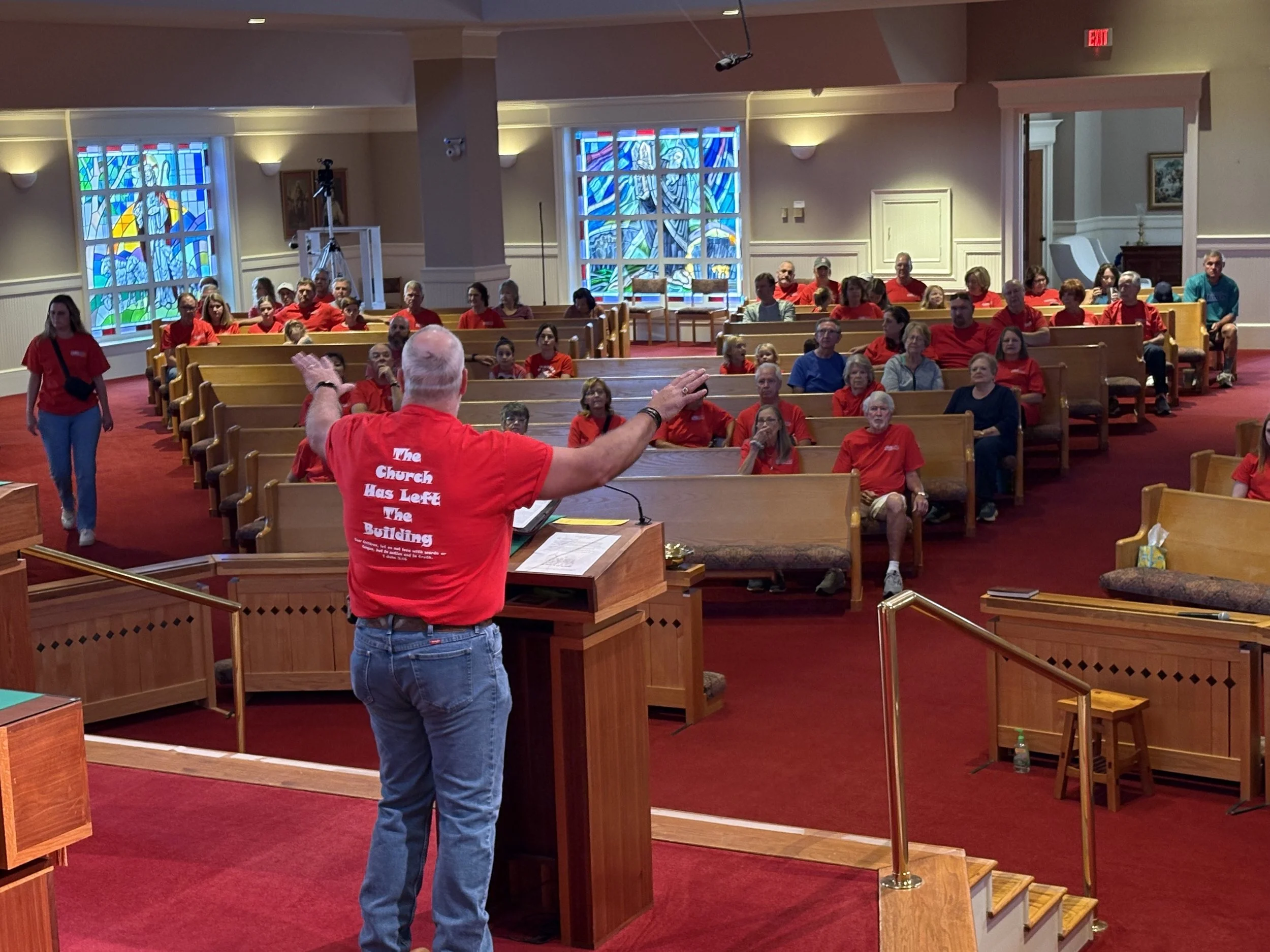 A man with a beard wearing a red shirt and jeans is speaking to a congregation inside a church. The congregation, dressed mainly in red, is seated on wooden pews. The church has stained glass windows and wooden accents.