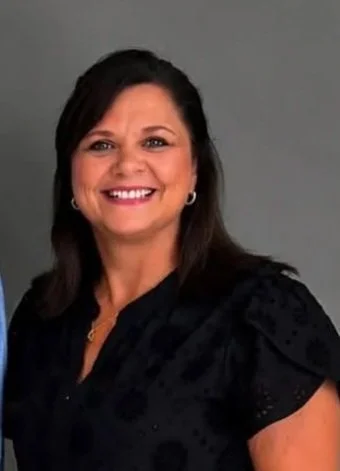 A smiling woman with dark hair, wearing a black top, standing against a gray background.