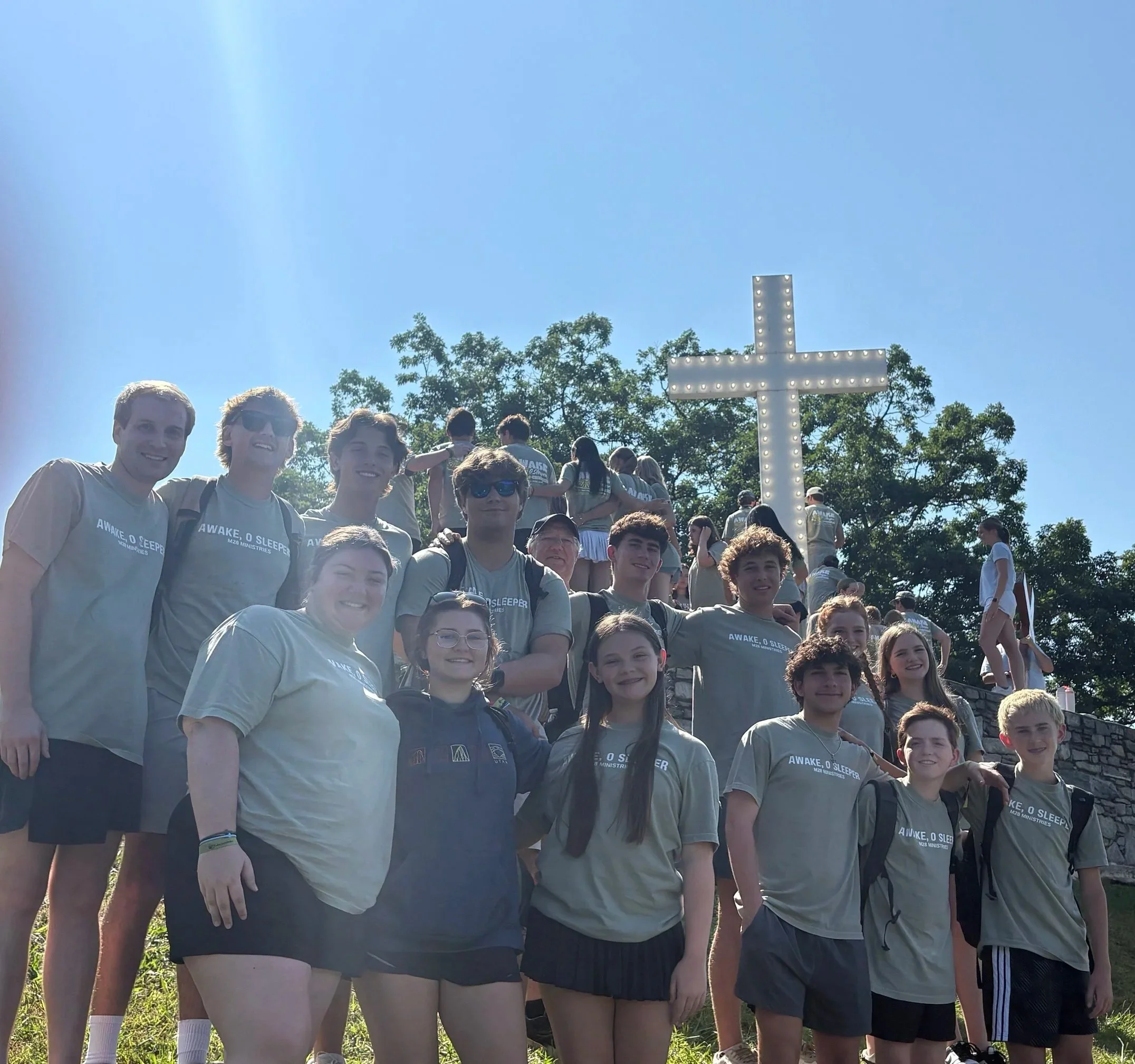 A group of young people outdoors on a sunny day, some wearing matching gray T-shirts that read 'AWAKE, O SLEEPER' and 'M20 MINISTRIES,' with trees in the background and a large illuminated cross beside them.