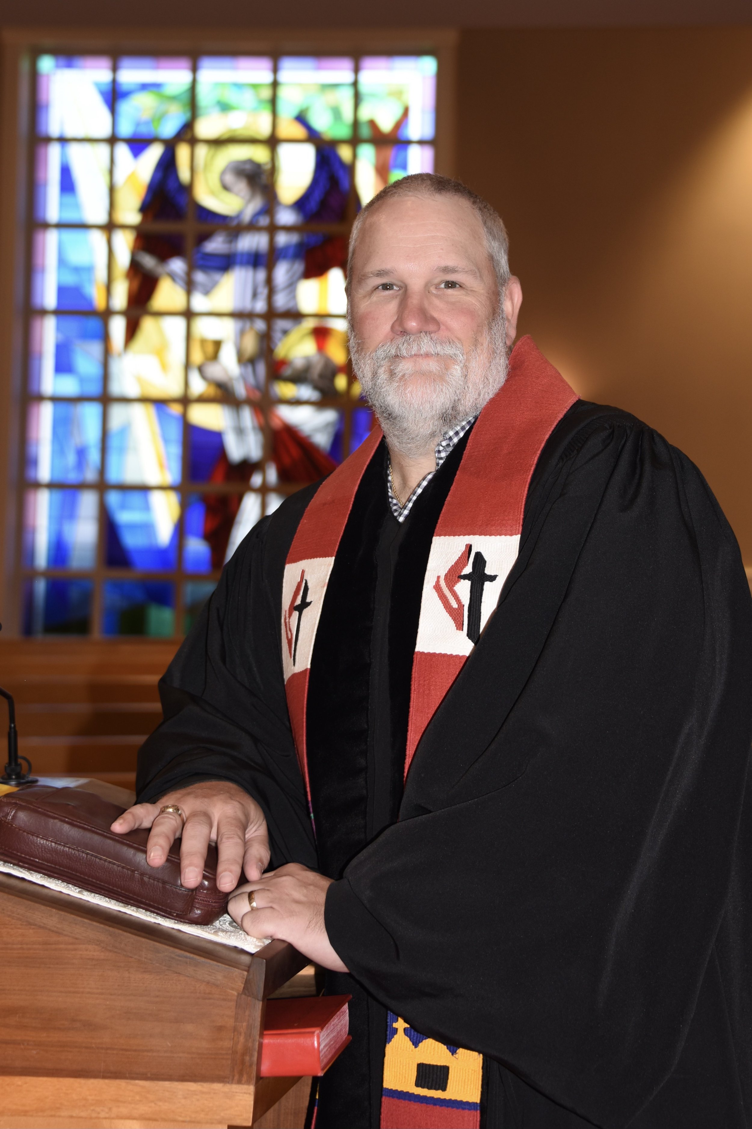 A man with a beard and mustache in a black robe and red stole, sitting at a wooden desk, with a stained glass window of Jesus Christ behind him.