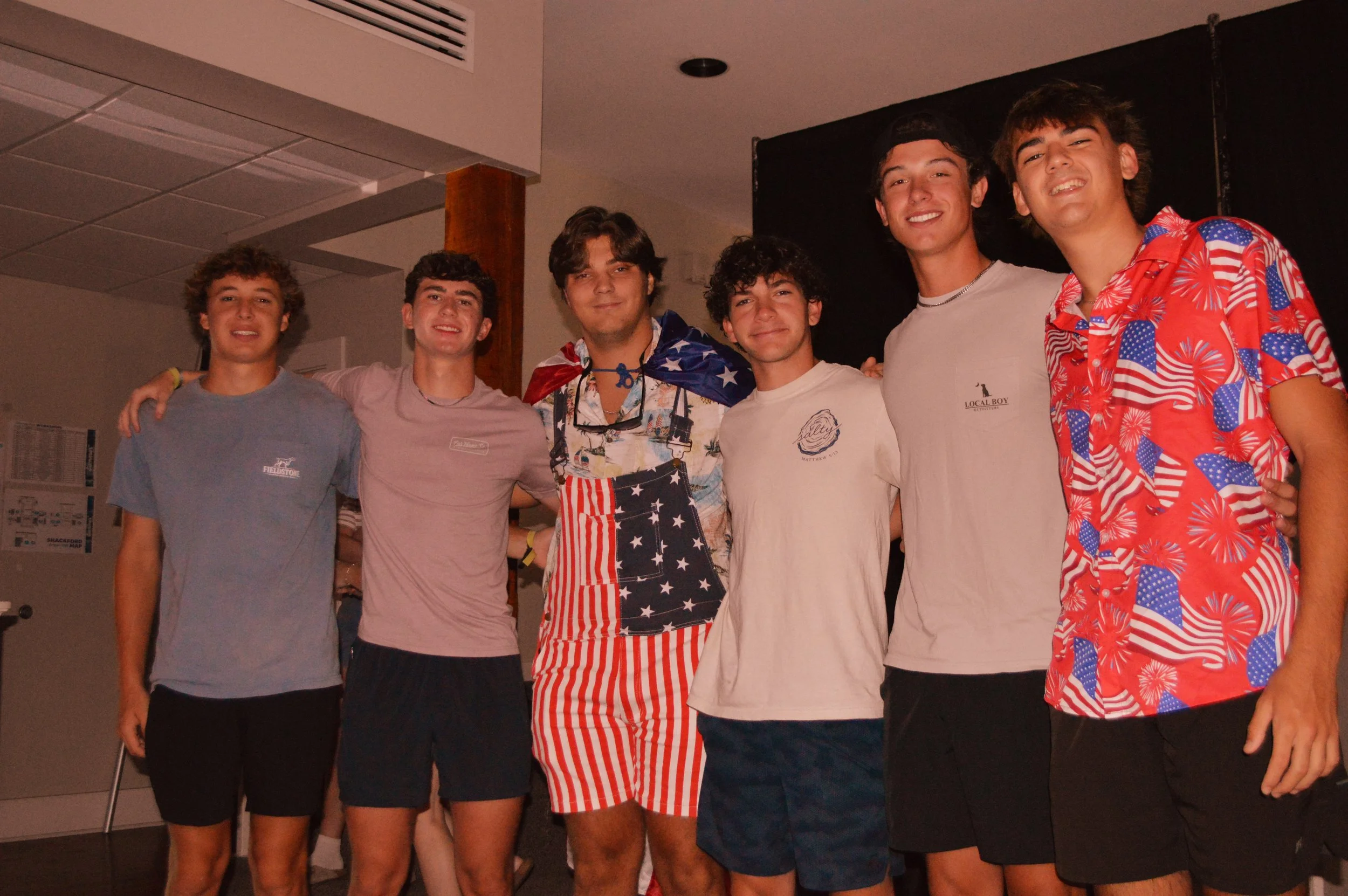 Seven young men standing close together at a party or gathering, dressed casually, with some wearing patriotic-themed clothing and accessories, smiling and posing for the photo.