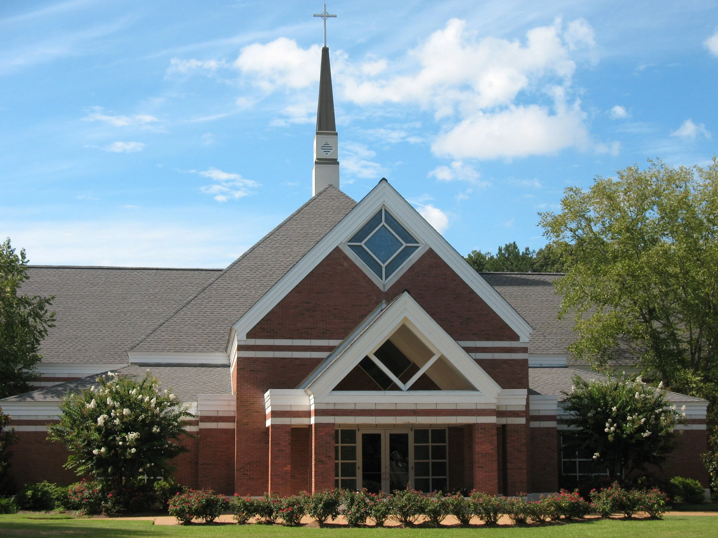 Front view of a church with a steep roof, brick walls, a tall steeple with a cross, decorative window, surrounded by greenery and flowering bushes, under a blue sky with scattered clouds.