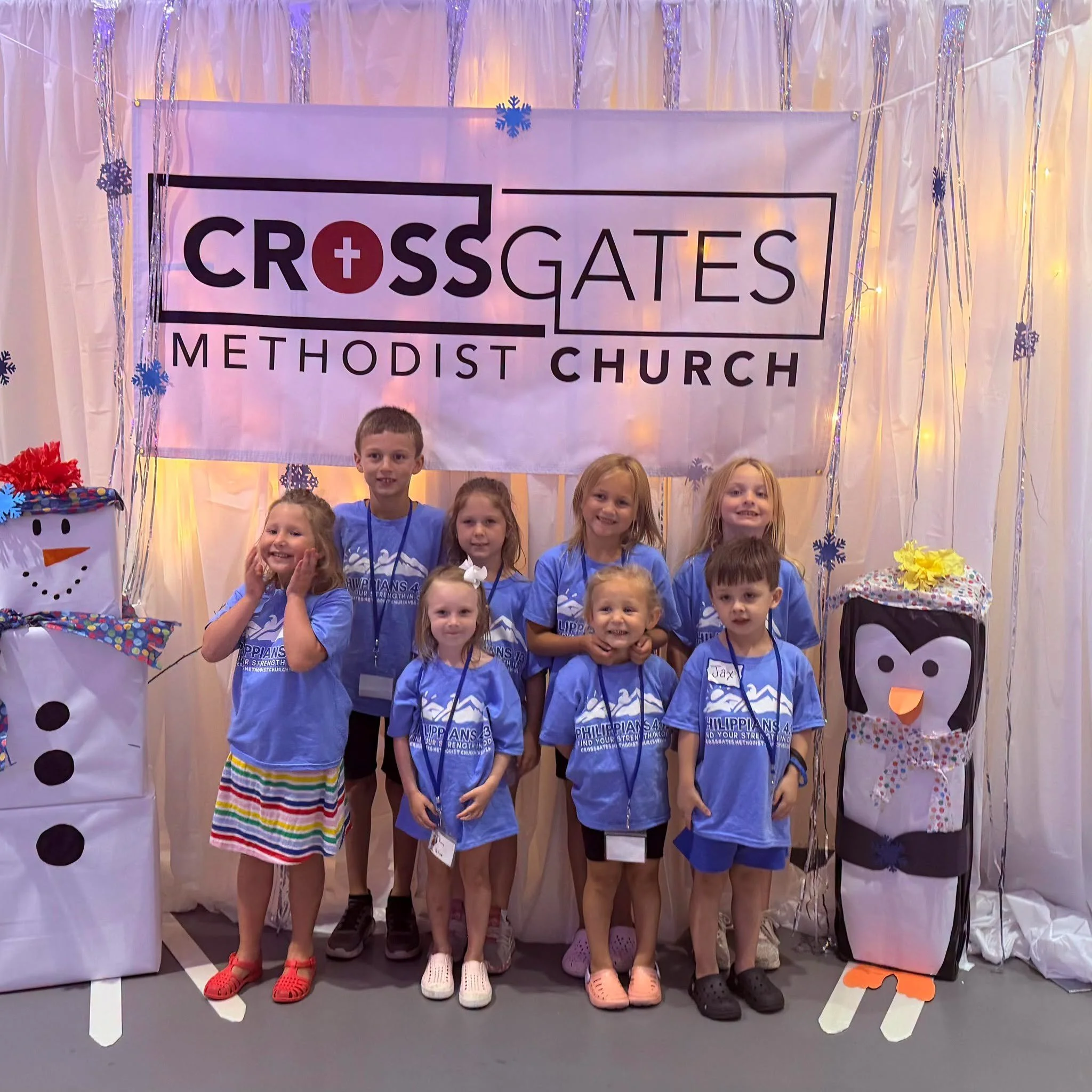 Group of children standing in front of a decorated backdrop with a banner that reads 'Crossgates Methodist Church'. The backdrop is adorned with snowflakes and string lights, and the children are wearing matching blue T-shirts with a logo and text.