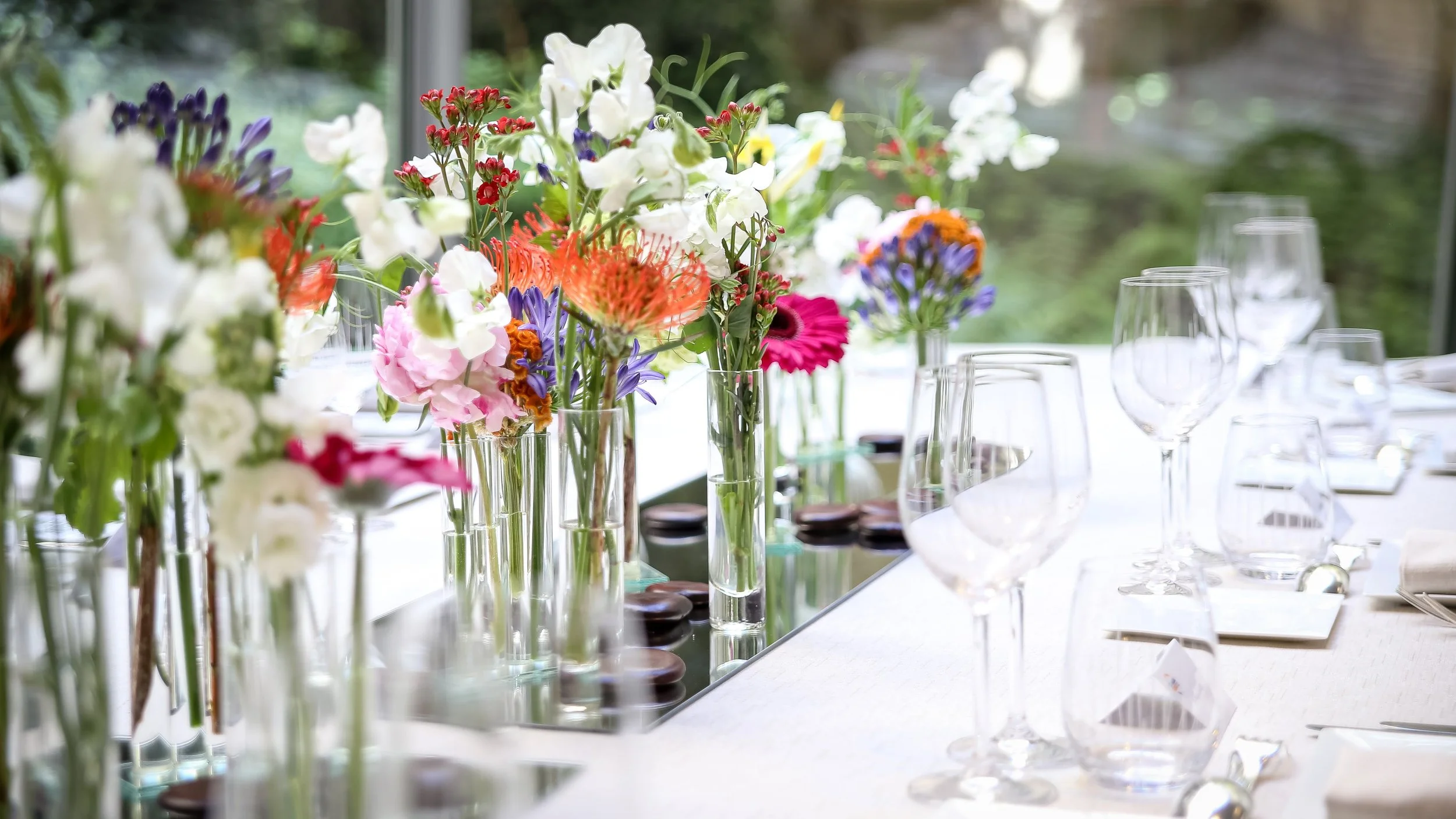 Vase de fleurs colorées sur une table, probablement pour une décoration d'événement ou un repas, avec des verres et couverts en arrière-plan.