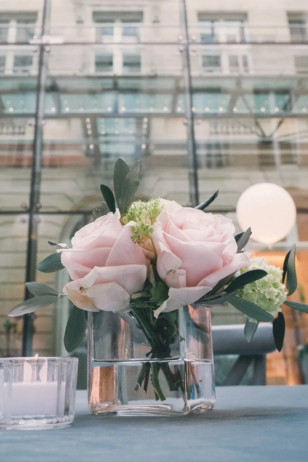 Bouquet de roses pâles dans un vase en verre, placé sur une table avec une bougie à côté, dans un intérieur moderne avec un mur en verre en arrière-plan.