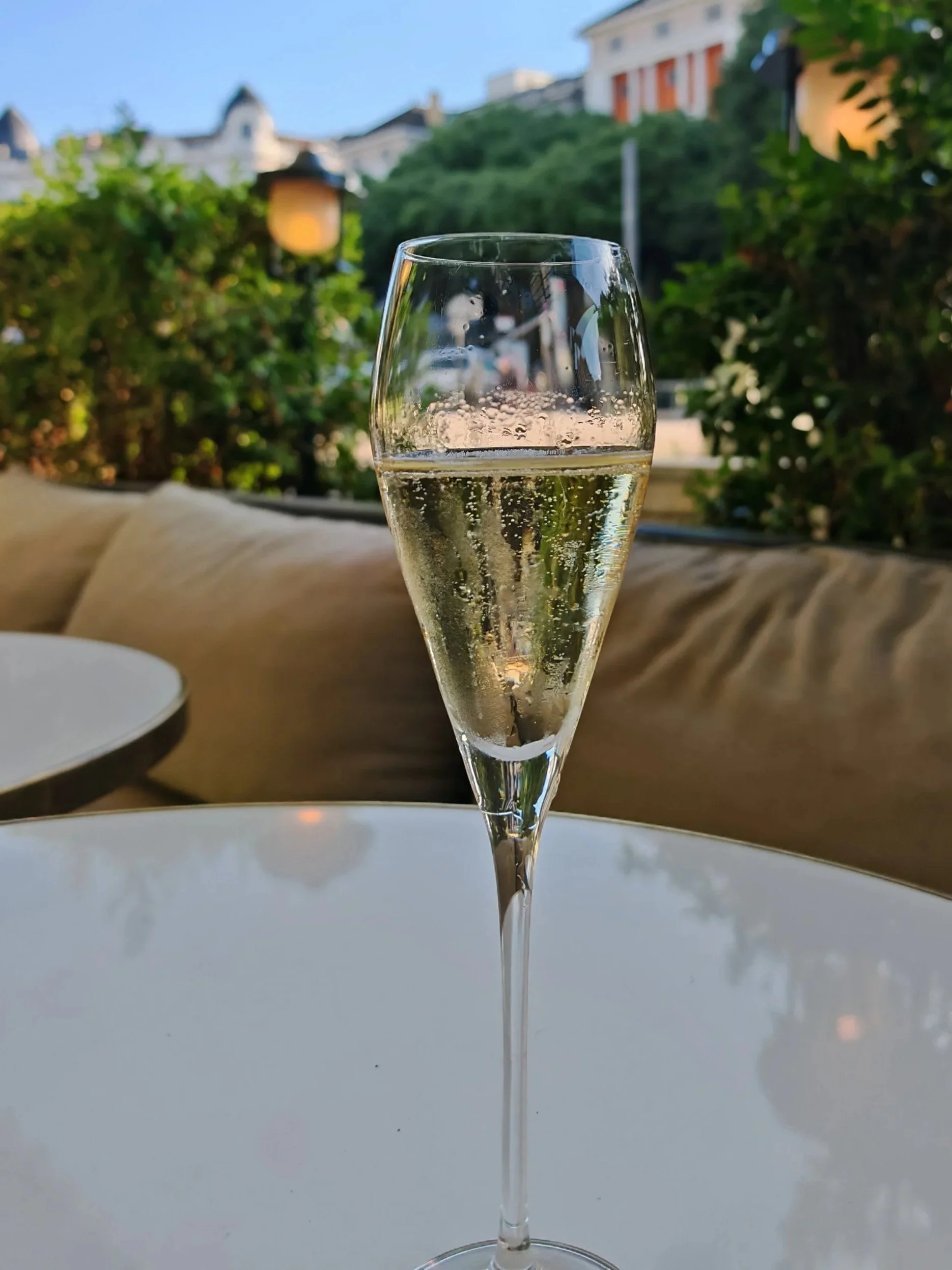 Verre de champagne rempli, placé sur une table blanche, avec un fond de verdure et des maisons en arrière-plan par une journée ensoleillée.
