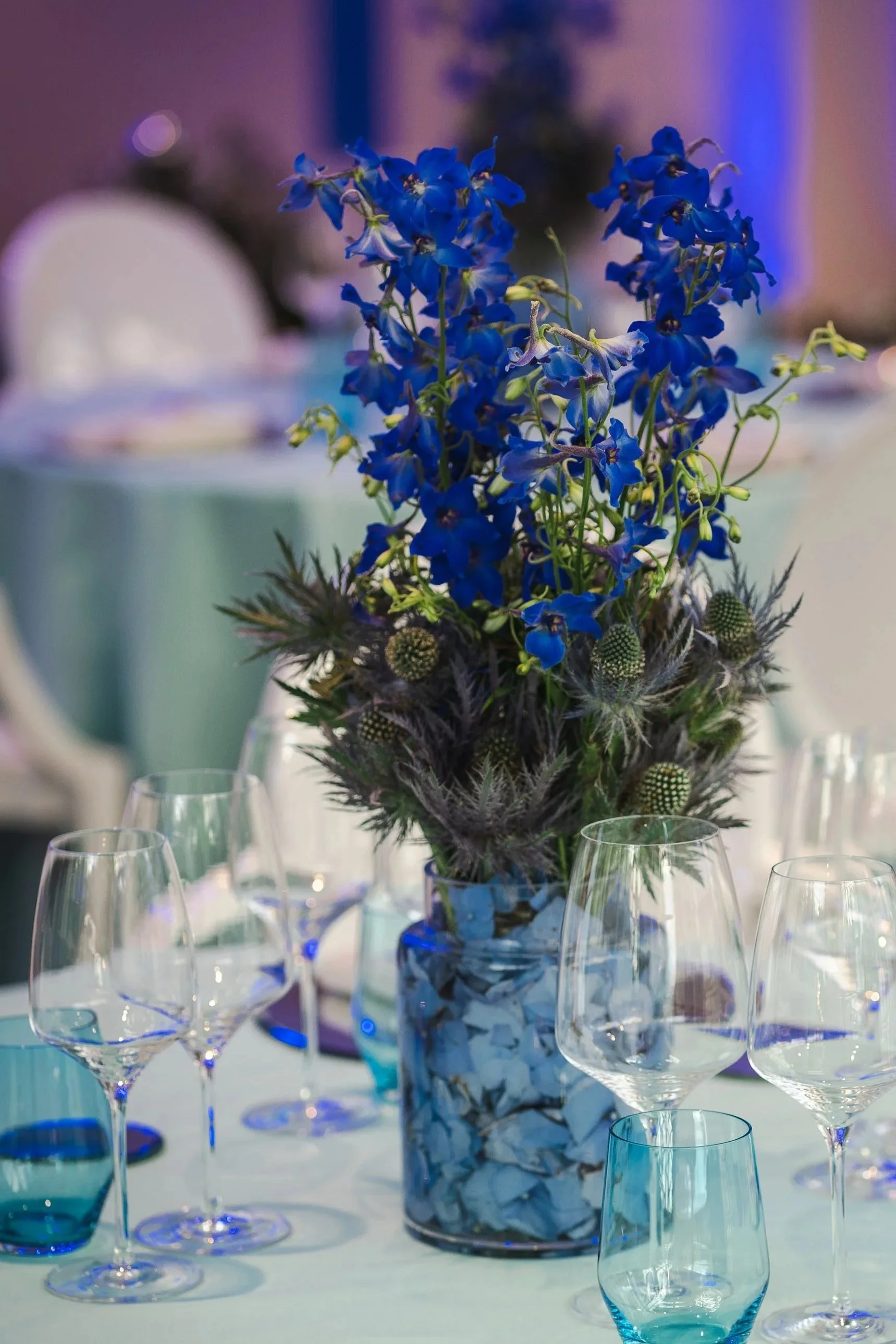 Un bouquet de fleurs bleues dans un vase, entouré de verres à vin sur une table, dans un décor de fête ou de réception.