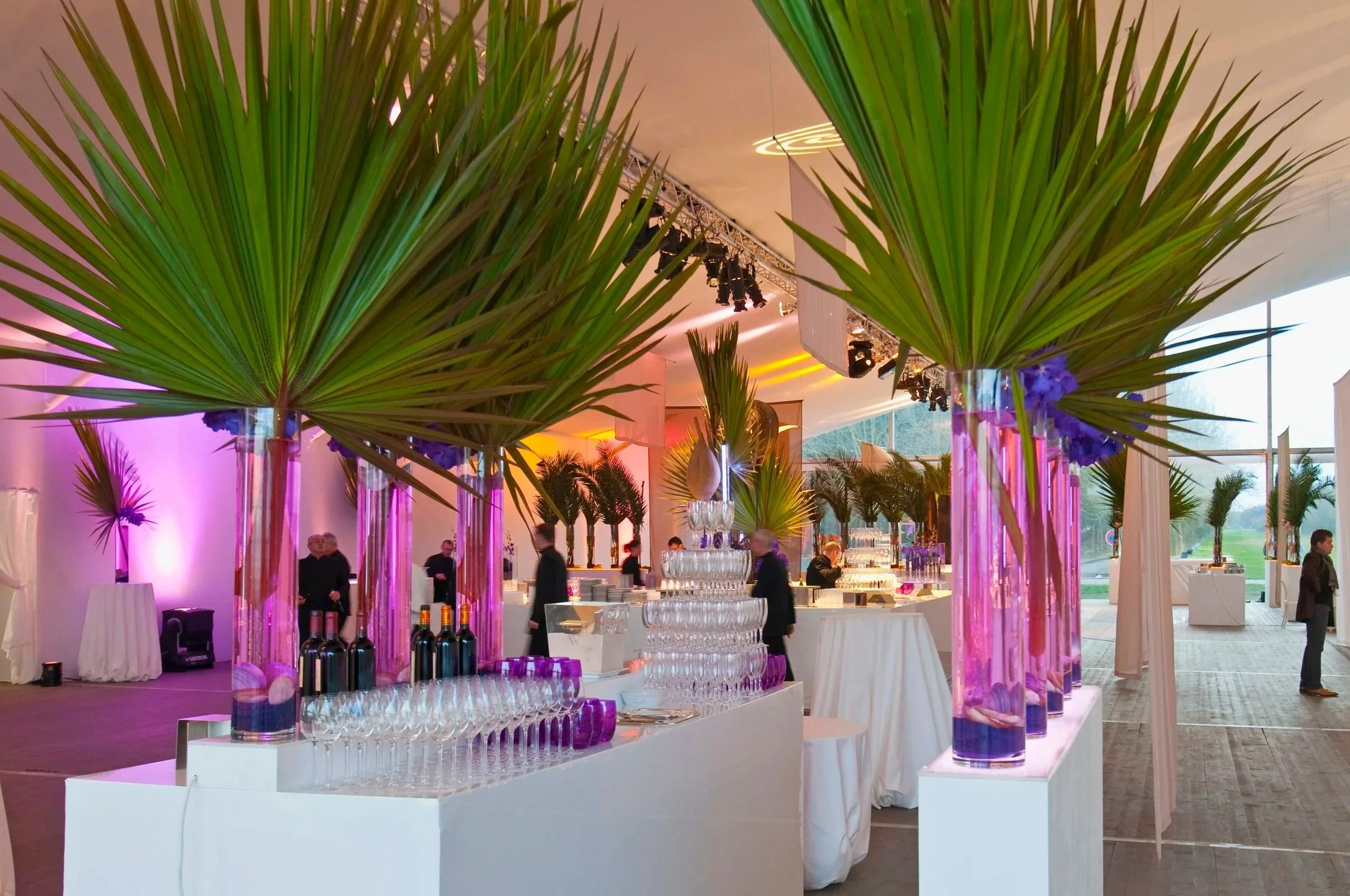 Décor de banquet avec grandes plantes en vases en verre coloré violet, ambiance élégante avec lumière tamisée, personne au bar, plafond avec éclairage moderne.