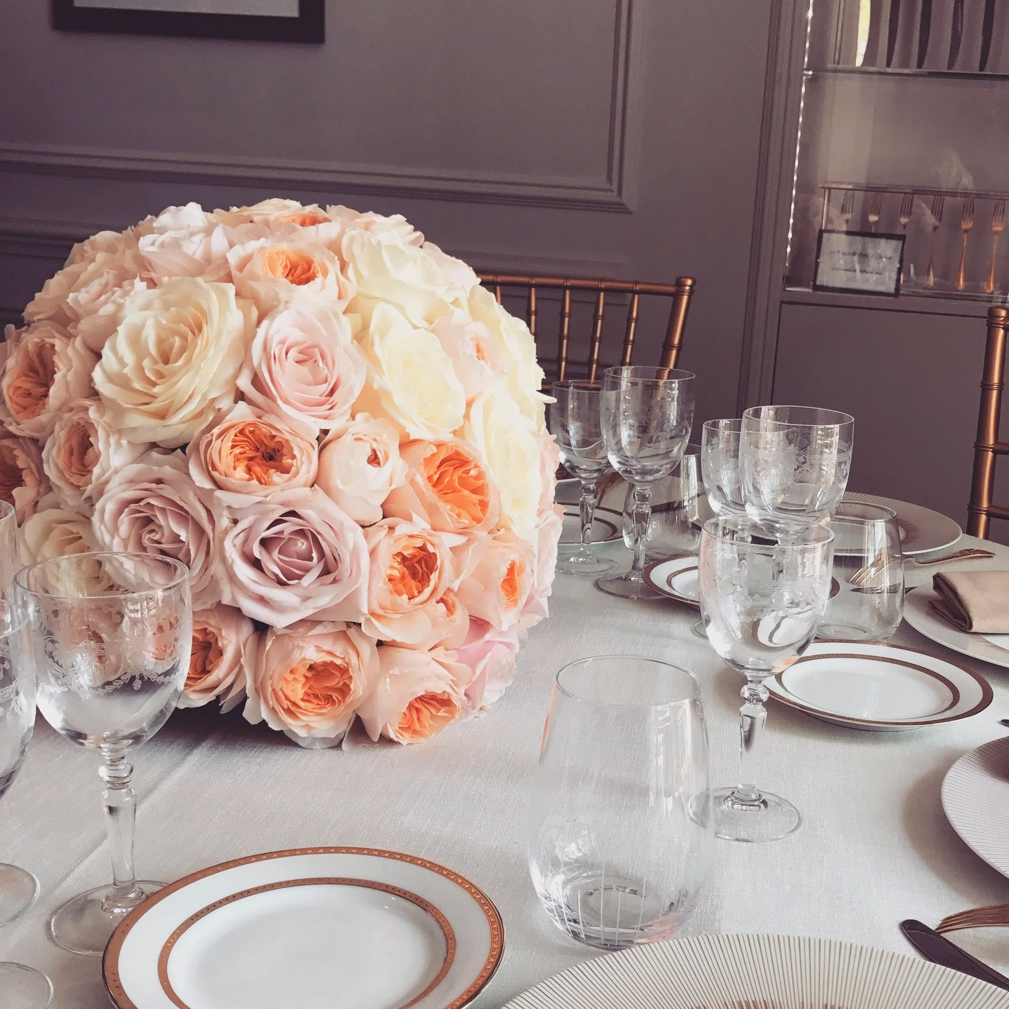 Un centre de table composé de nombreuses roses aux teintes de blanc, beige et pêche sur une table de dîner élégante, entouré de verres à vin transparents et de vaisselle fine dans une salle décorée de gris avec des chaises en métal doré.