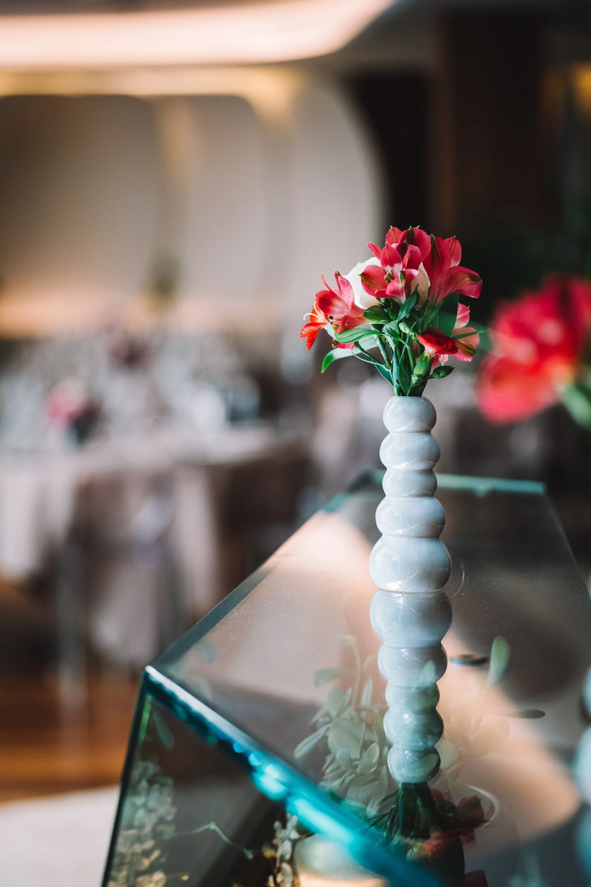 Un bouquet de fleurs roses et blanches dans un vase blanc posé sur une surface en miroir. La photo est prise à l'intérieur, avec un fond flou.