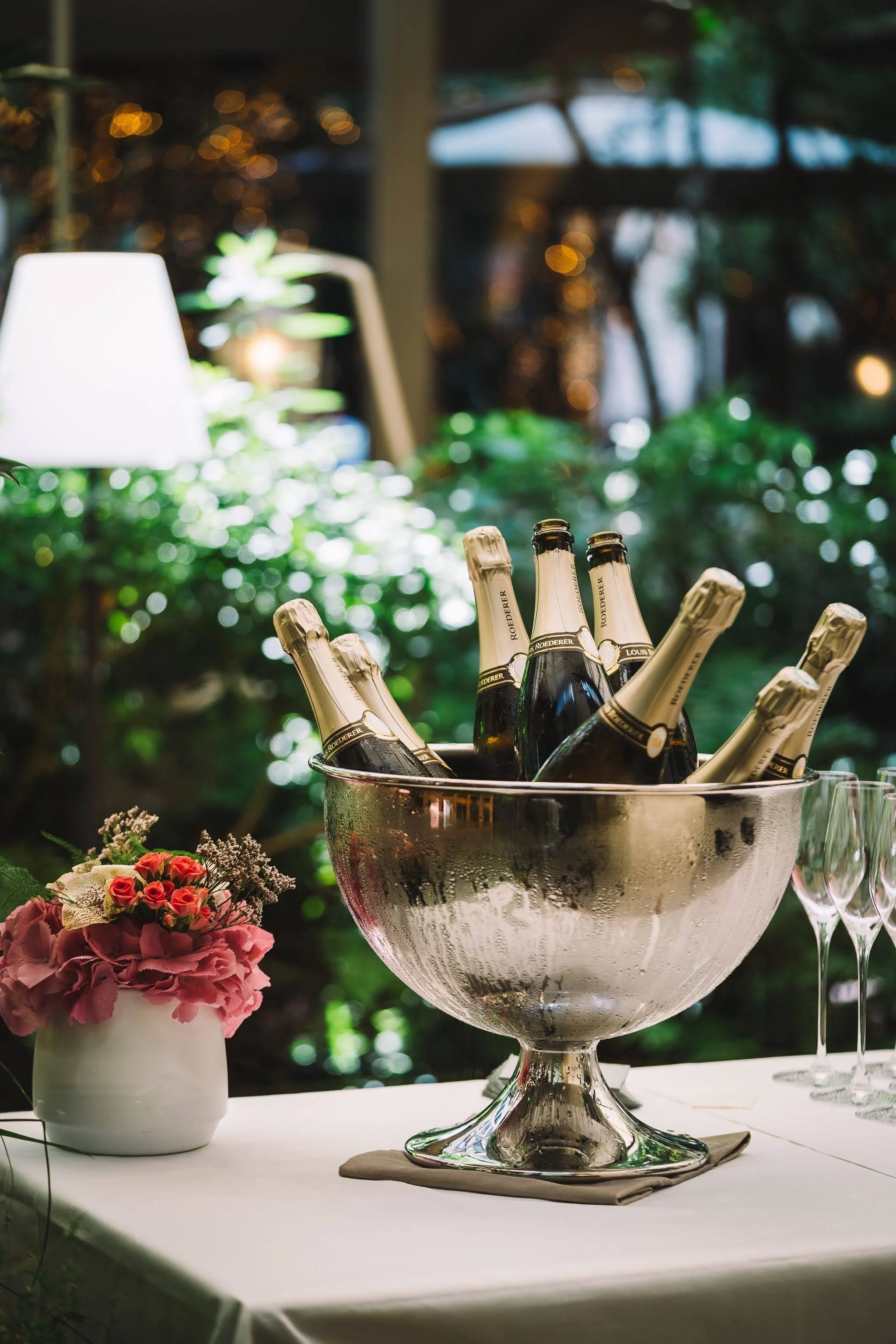 Verre à cocktail avec bouteilles de champagne dans un bol en argent, décor floral avec un vase de roses roses sur une table, ambiance élégante et festive dans un environnement extérieur ou intérieur bien éclairé.
