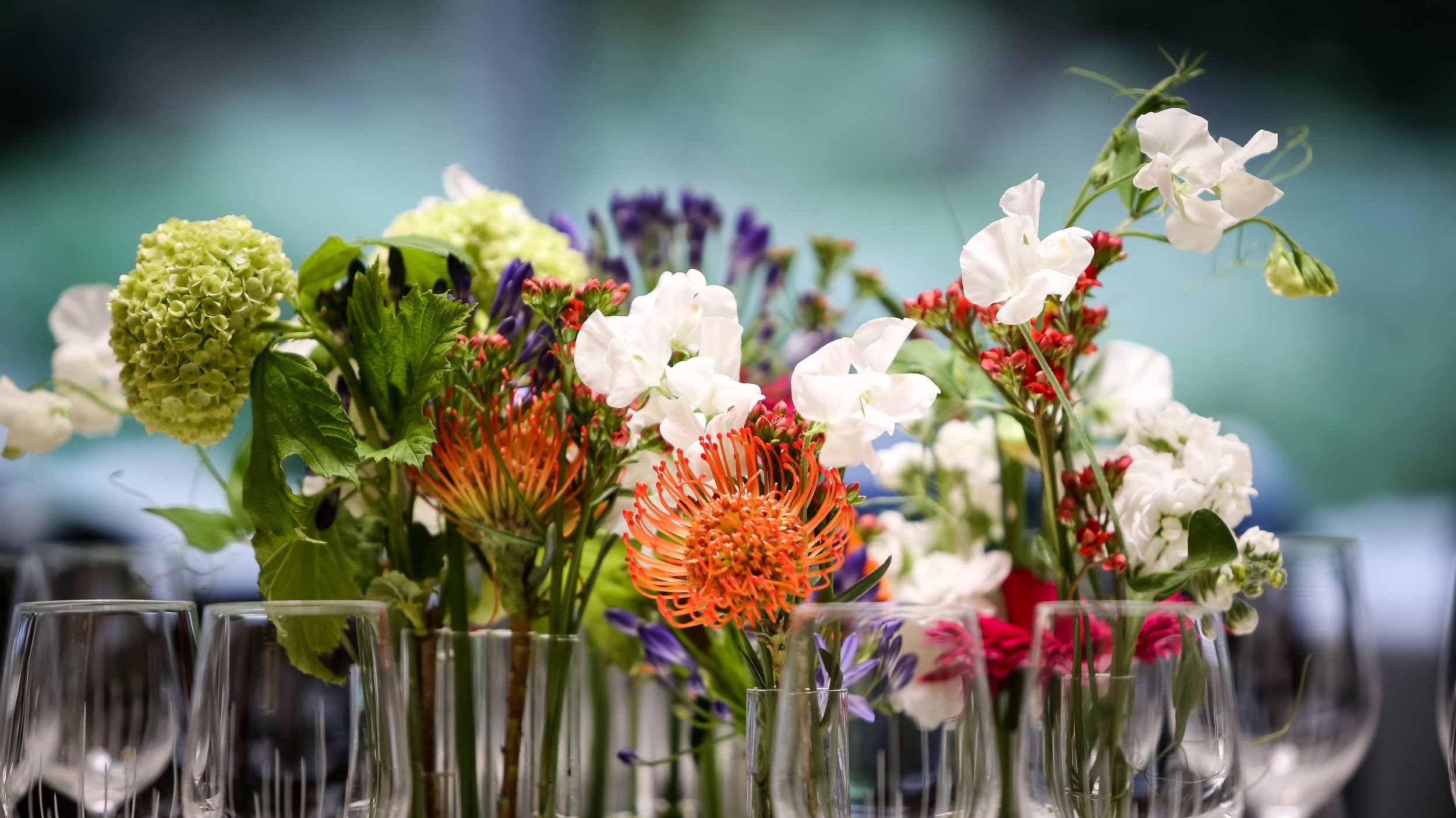 Un arrangement de fleurs colorées dans des vases en verre, placé sur une table, avec des verres à vin en première plan.
