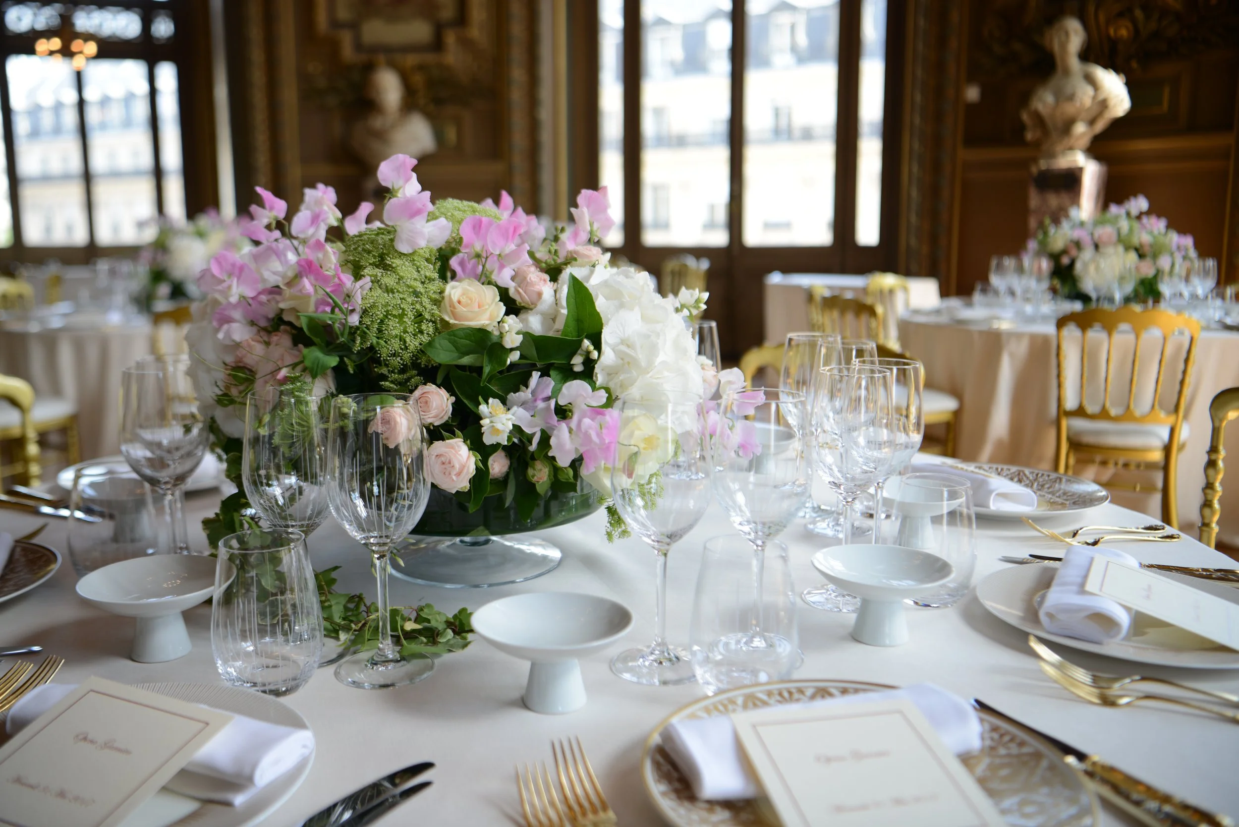 Table élégamment dressée avec un grand centre de fleurs combinant roses, hortensias et feuillage vert. Autour, des assiettes, verres, couverts en or, et des menus sur des napperons blancs sont disposés, dans une salle ornée de statues et de fenêtres 