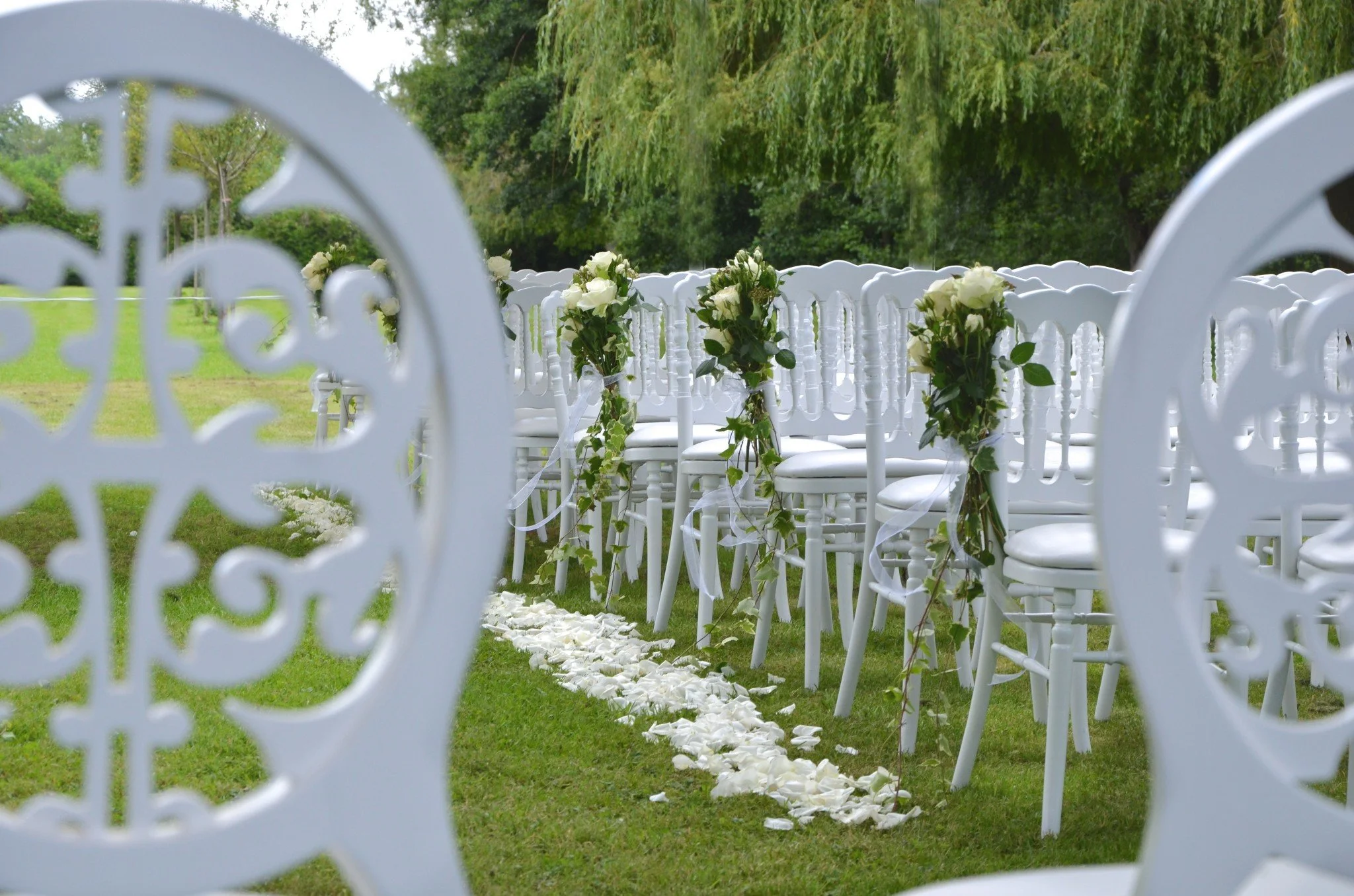 Chaises blanches décorées de bouquets de roses blanches, disposées pour une cérémonie de mariage en plein air. La scène est encadrée par une grille en métal blanc à motif floral.