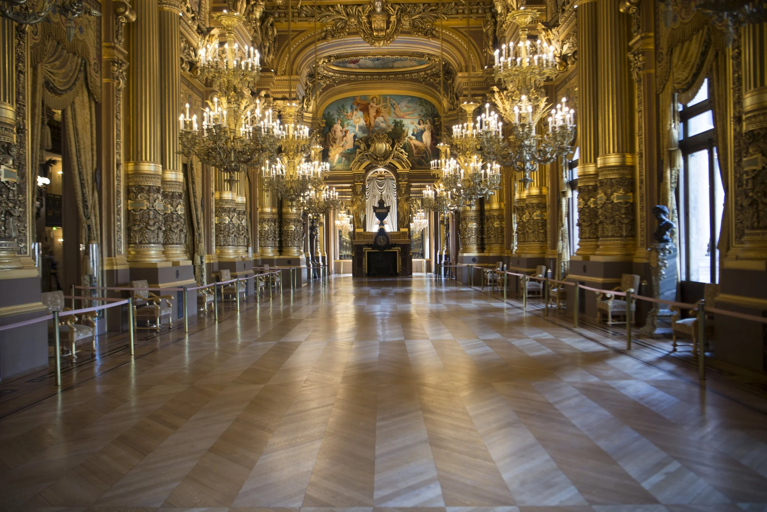 Salle grandiose avec chandeliers en cristal dorés, murs ornés de moulures et de dorures, colonnes décoratives, tableaux peints et très grande fenêtre laissant entrer la lumière naturelle.