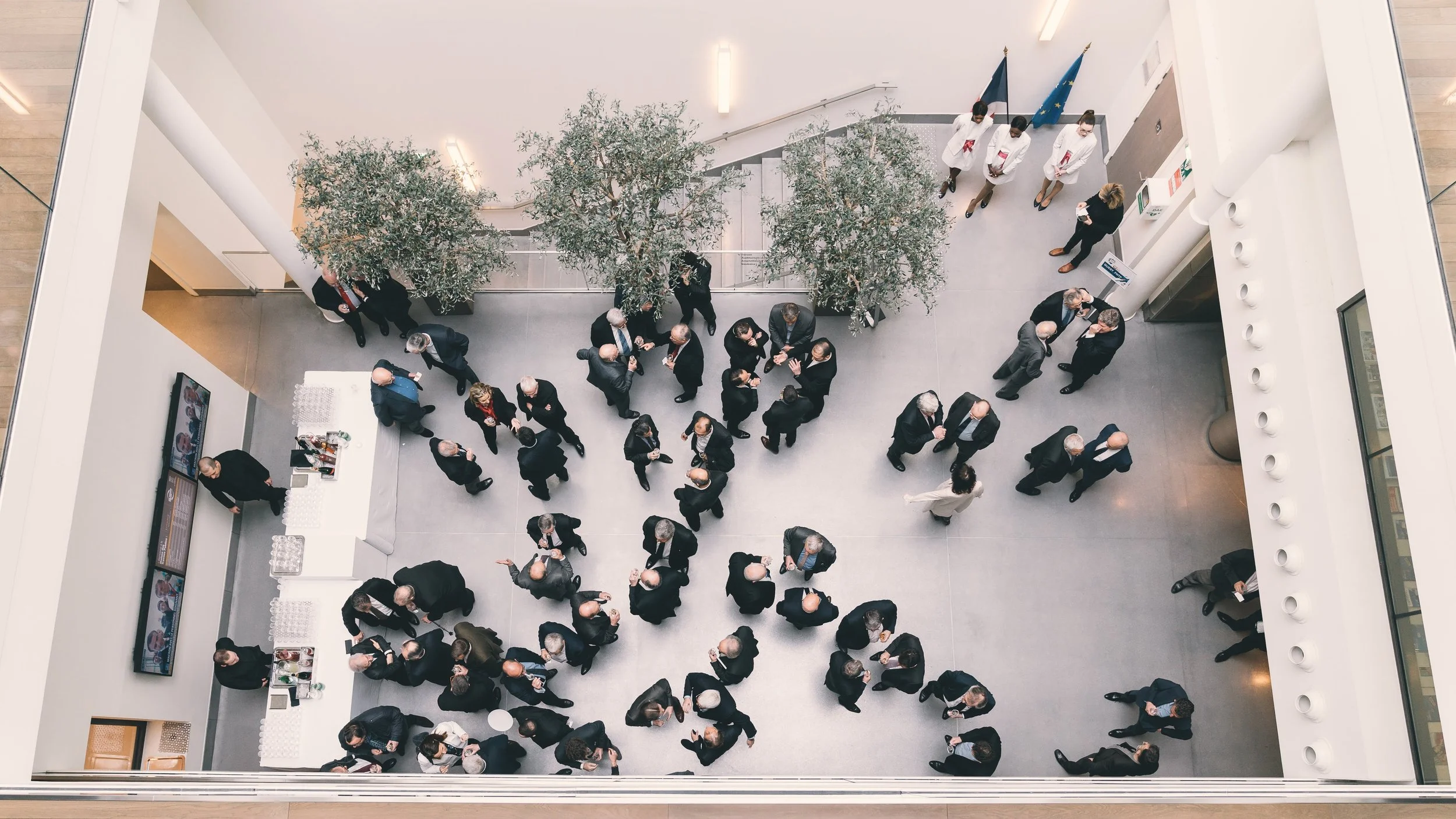 Une vue d'en haut d'une foule de personnes habillées en noir et blanc dans un espace intérieur moderne avec deux arbres au centre, entourées de murs blancs, une bannière avec des drapeaux en arrière-plan et un comptoir avec des boissons.