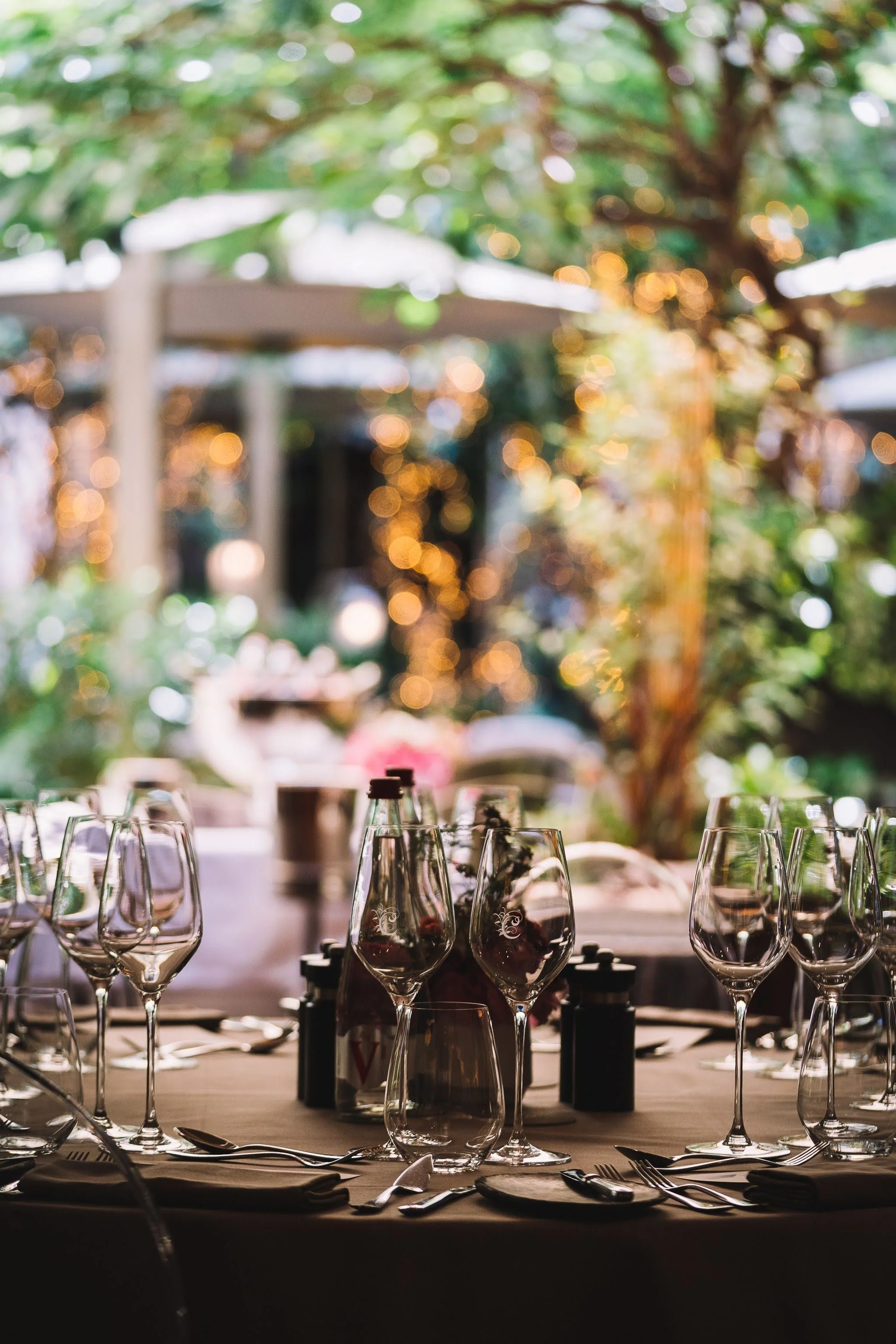 Une table de banquet élégante décorée de verres à vin, de bouteilles de vin, de vaisselle et de couverts, avec un fond flou de décorations lumineuses et de végétation, évoquant une ambiance de mariage ou réception en plein air.