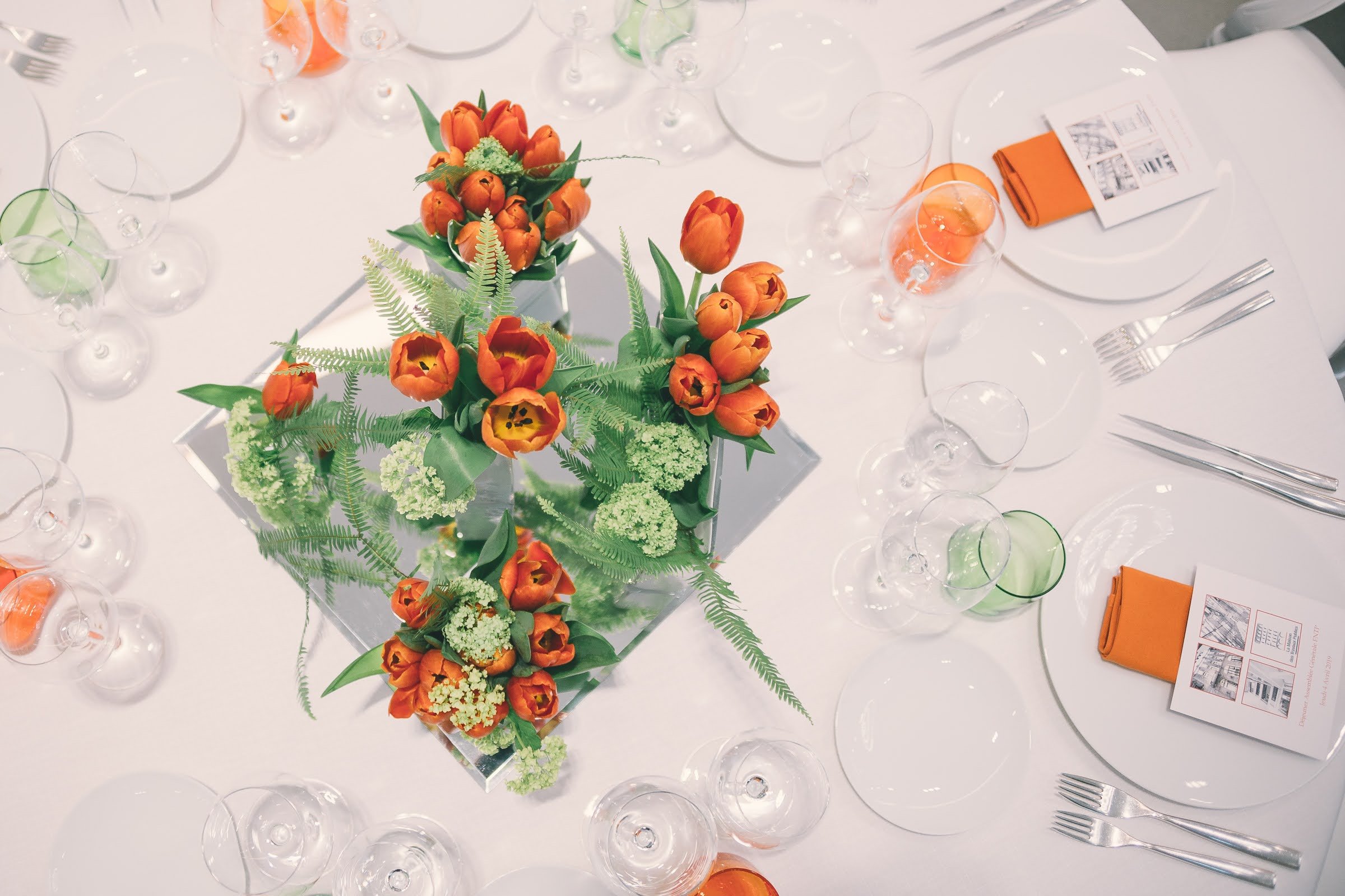 Centre de table avec des fleurs orange, comprenant des tulipes, dans un décor de table de dîner élégant avec des verres à vin, des couverts et des napperons orange.