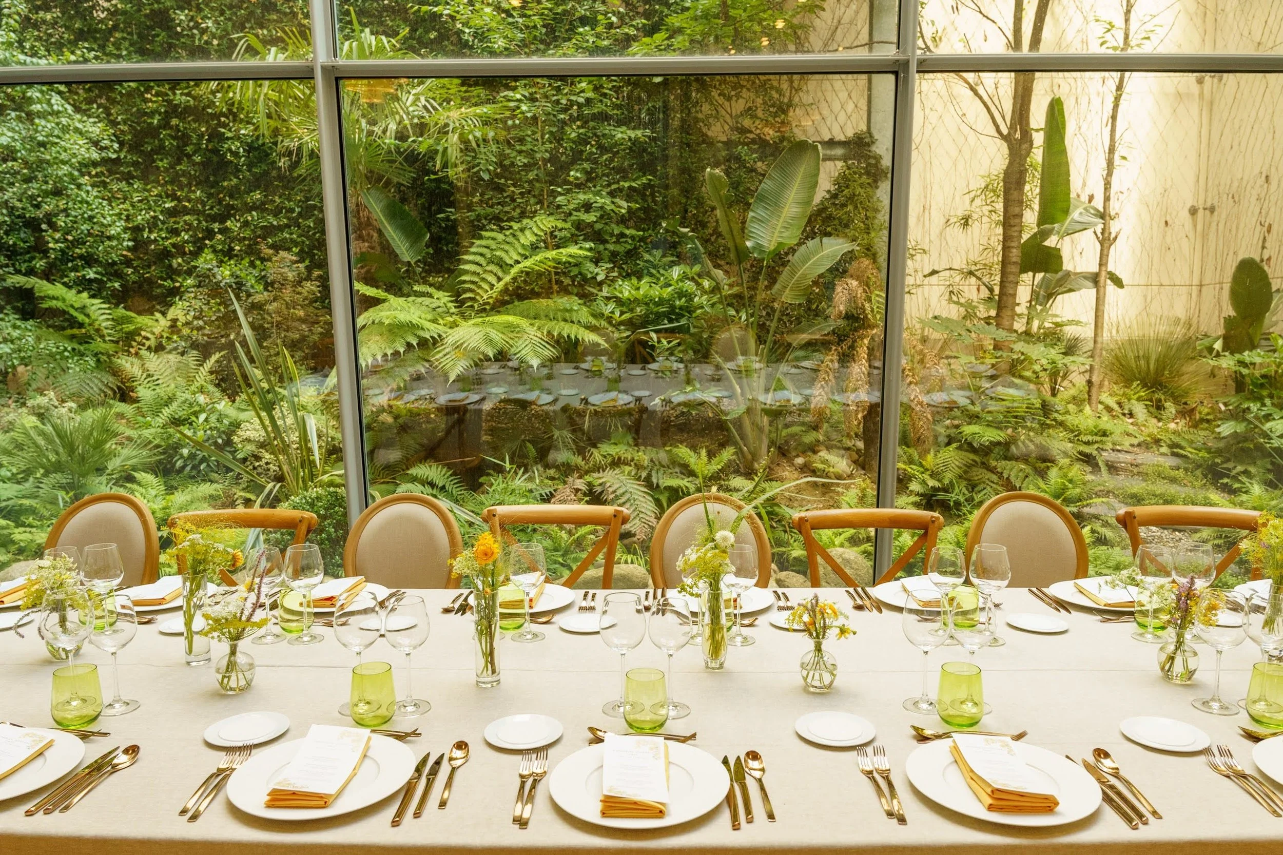 Une table dressée pour un repas avec des assiettes, des verres, des couverts et des petits bouquets de fleurs, vue à travers une grande fenêtre donnant sur un jardin avec beaucoup de verdure.