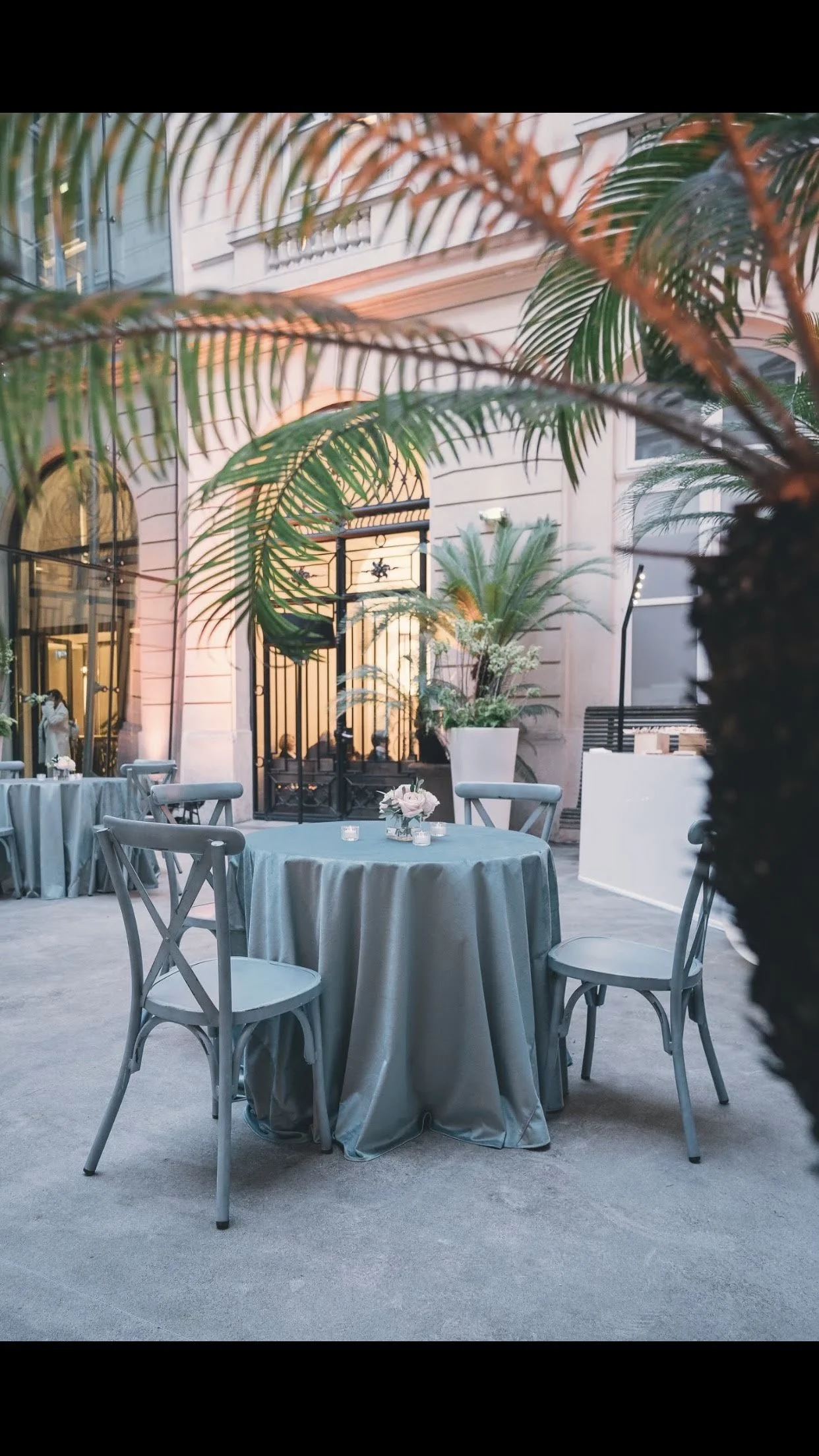 Une salle élégante avec des tables rondes recouvertes de nappes grises, décorées de fleurs et de bougies, entourée de chaises en bois, avec des plantes vertes et une architecture raffinée en arrière-plan.
