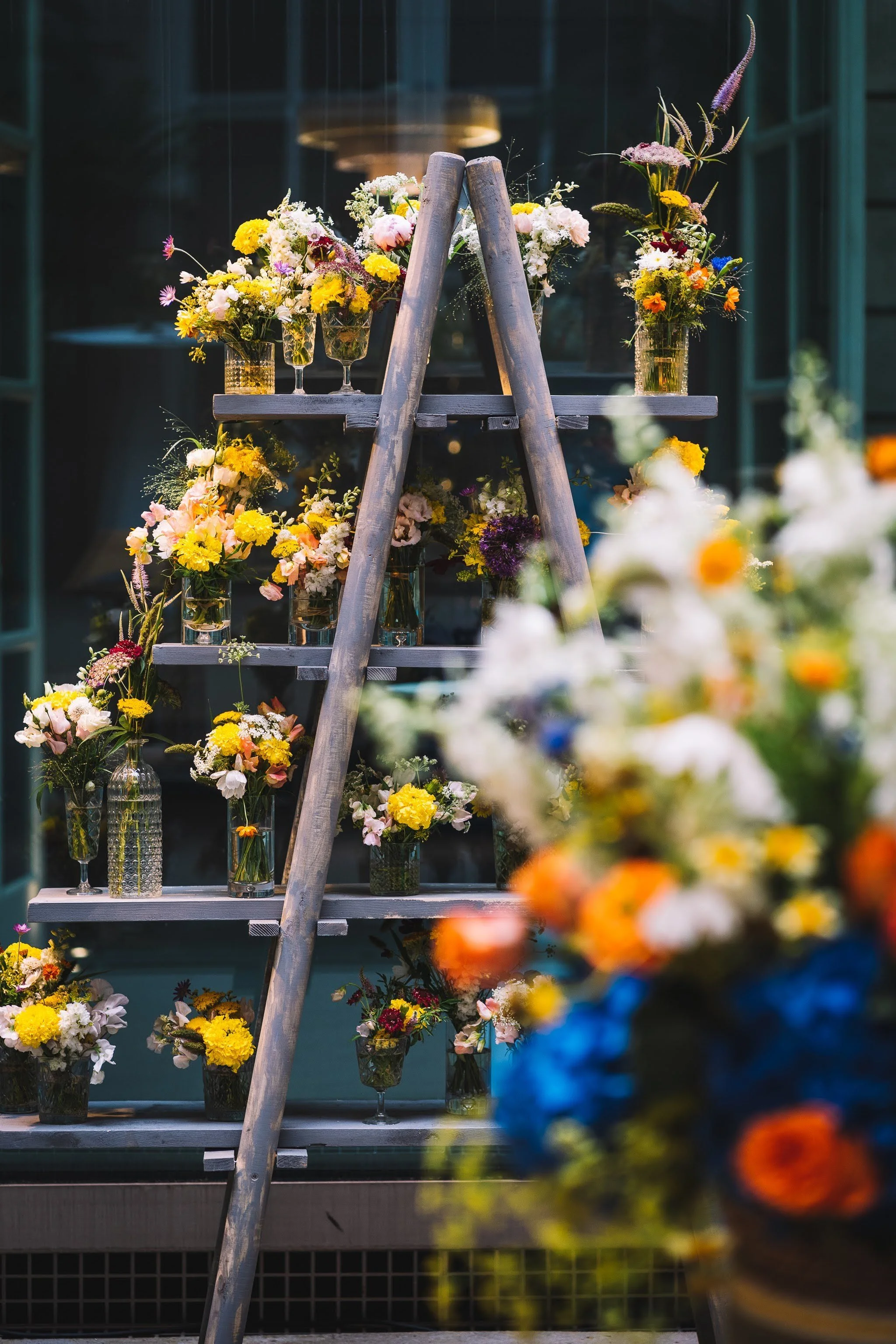 Arrangement floral avec différentes fleurs colorées dans des vases en verre sur une étagère en bois, avec un escabeau en bois à côté.