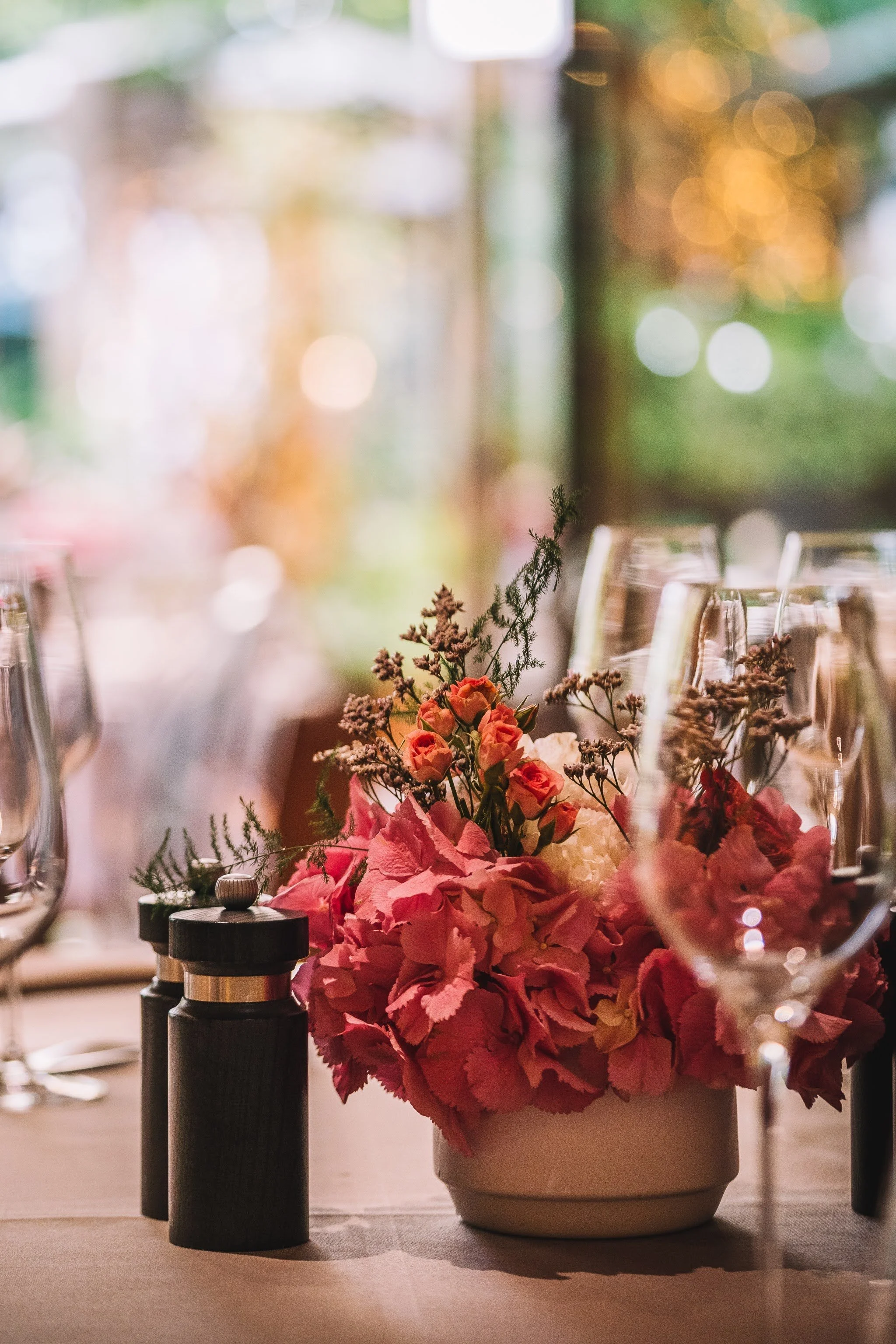 Un bouquet de fleurs roses et blanches dans un vase blanc, placé sur une table avec des bouteilles et des verres autour, ambiance chaleureuse et lumineuse.
