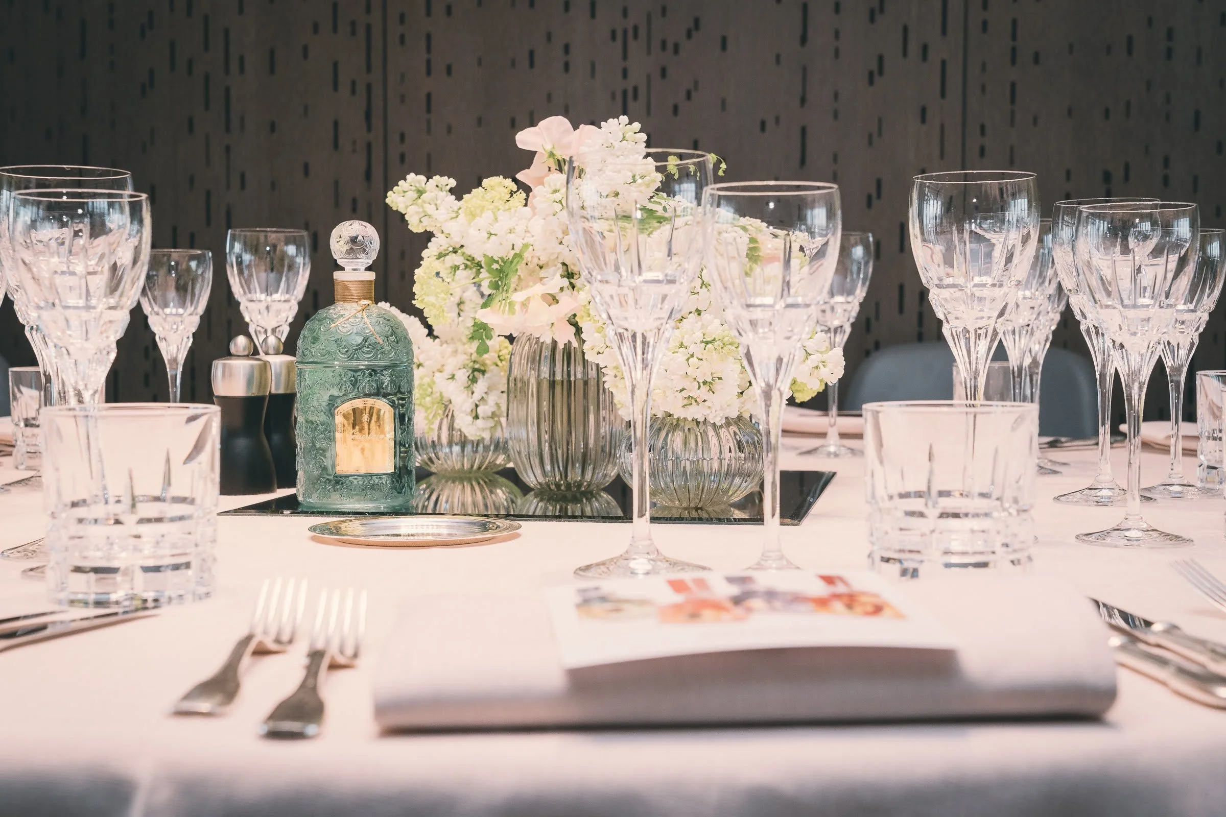 Table de banquet élégante avec des verres en cristal, un vase avec des fleurs blanches, un flacon de parfum vert, et des assiettes, dans un décor moderne.