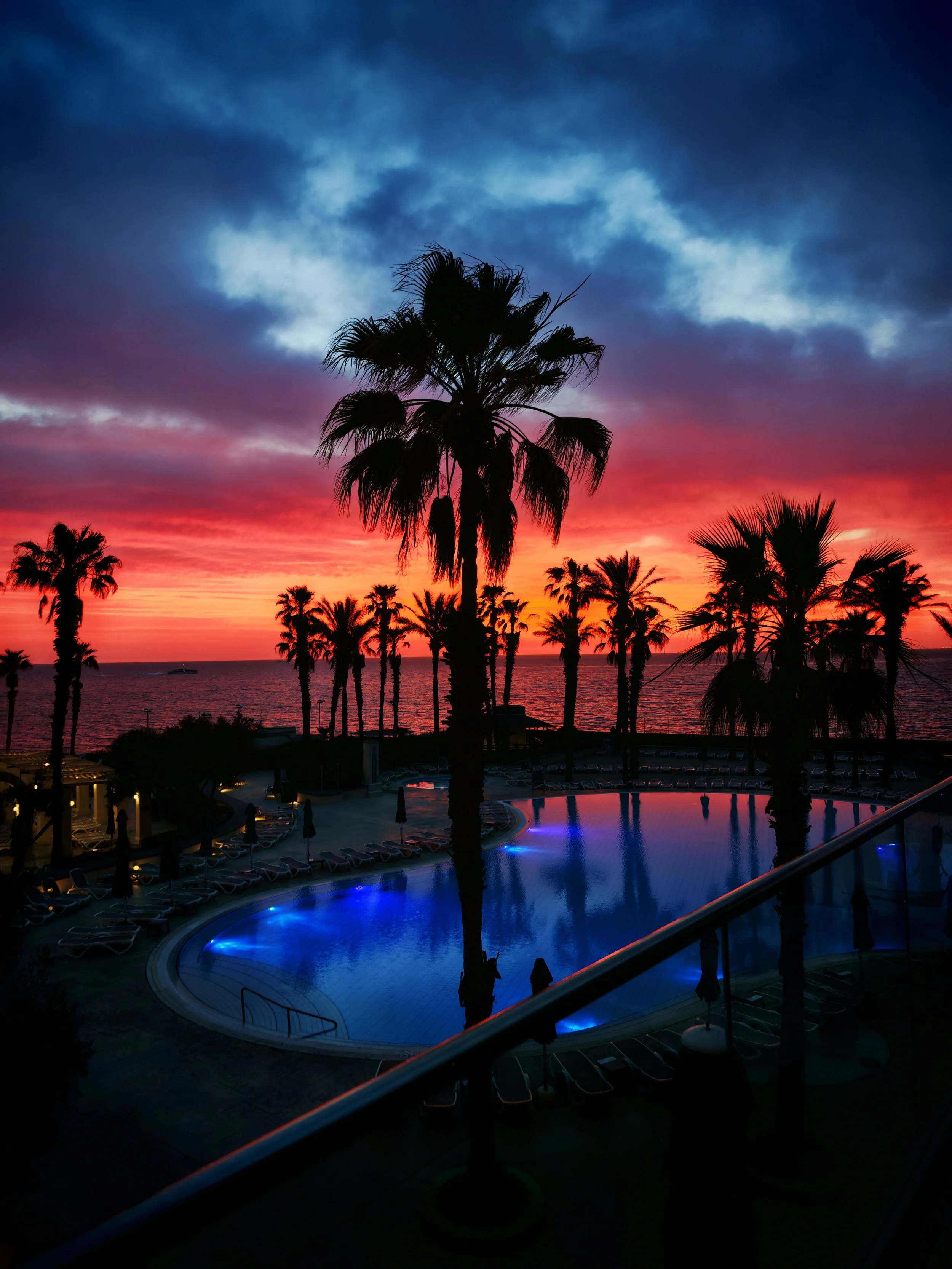 Couchers de soleil colorés derrière des palmiers sur la plage avec une piscine éclairée au premier plan.