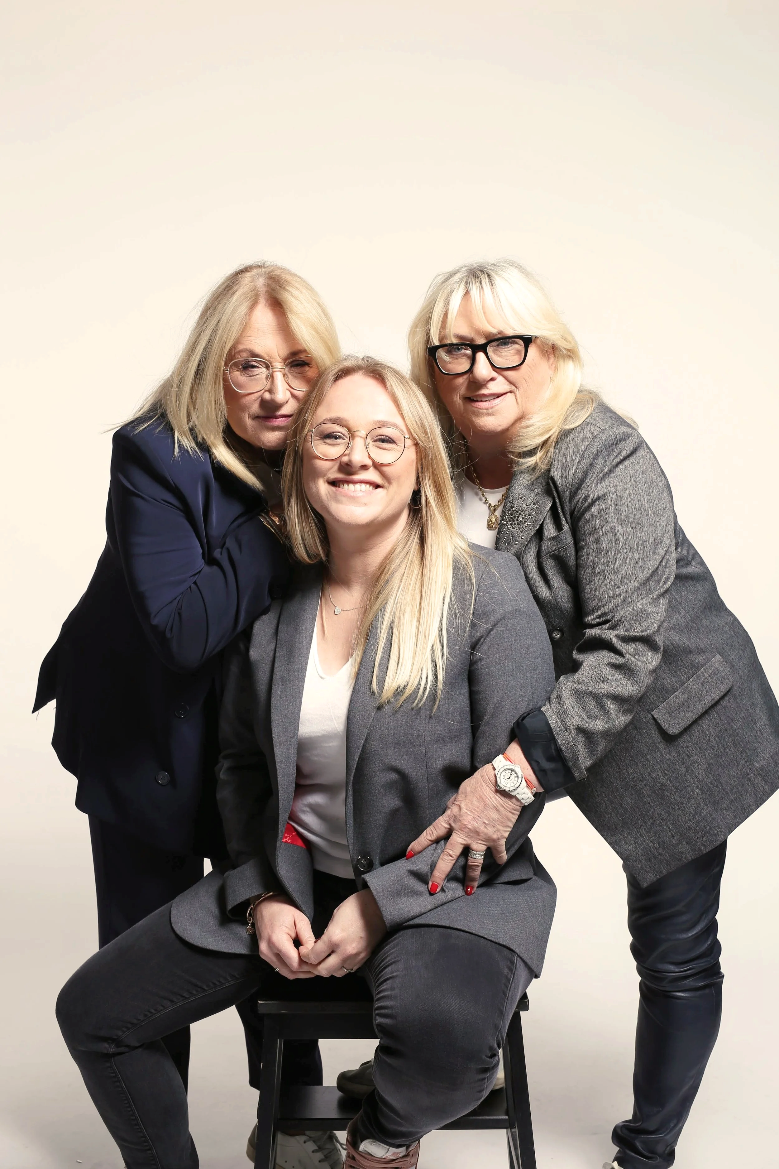 Trois femmes posant ensemble, souriantes, dans un fond neutre, habillées professionnellement, une assise ancienne en bois noir.