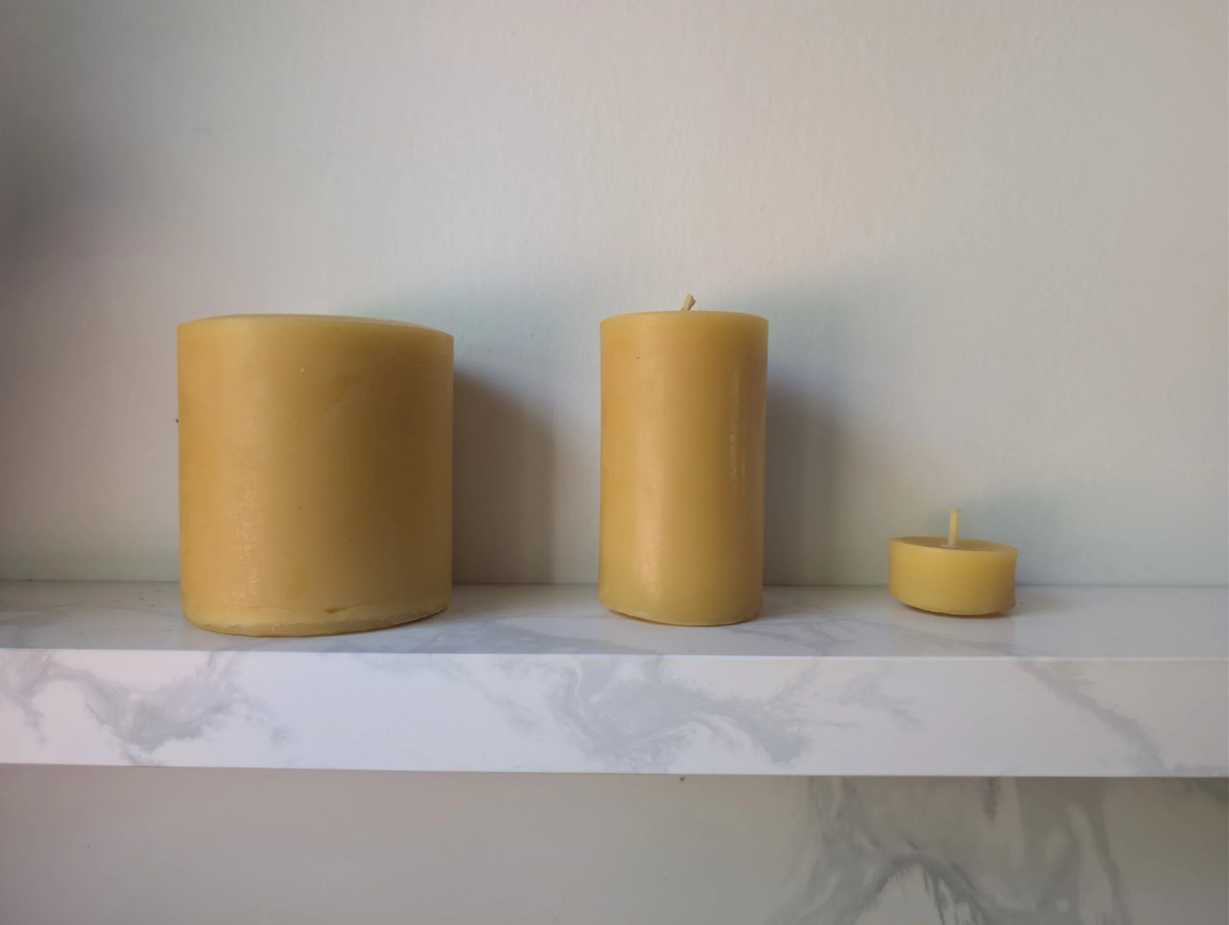 Three yellow candles of varying heights on a white shelf against a plain light-colored wall.