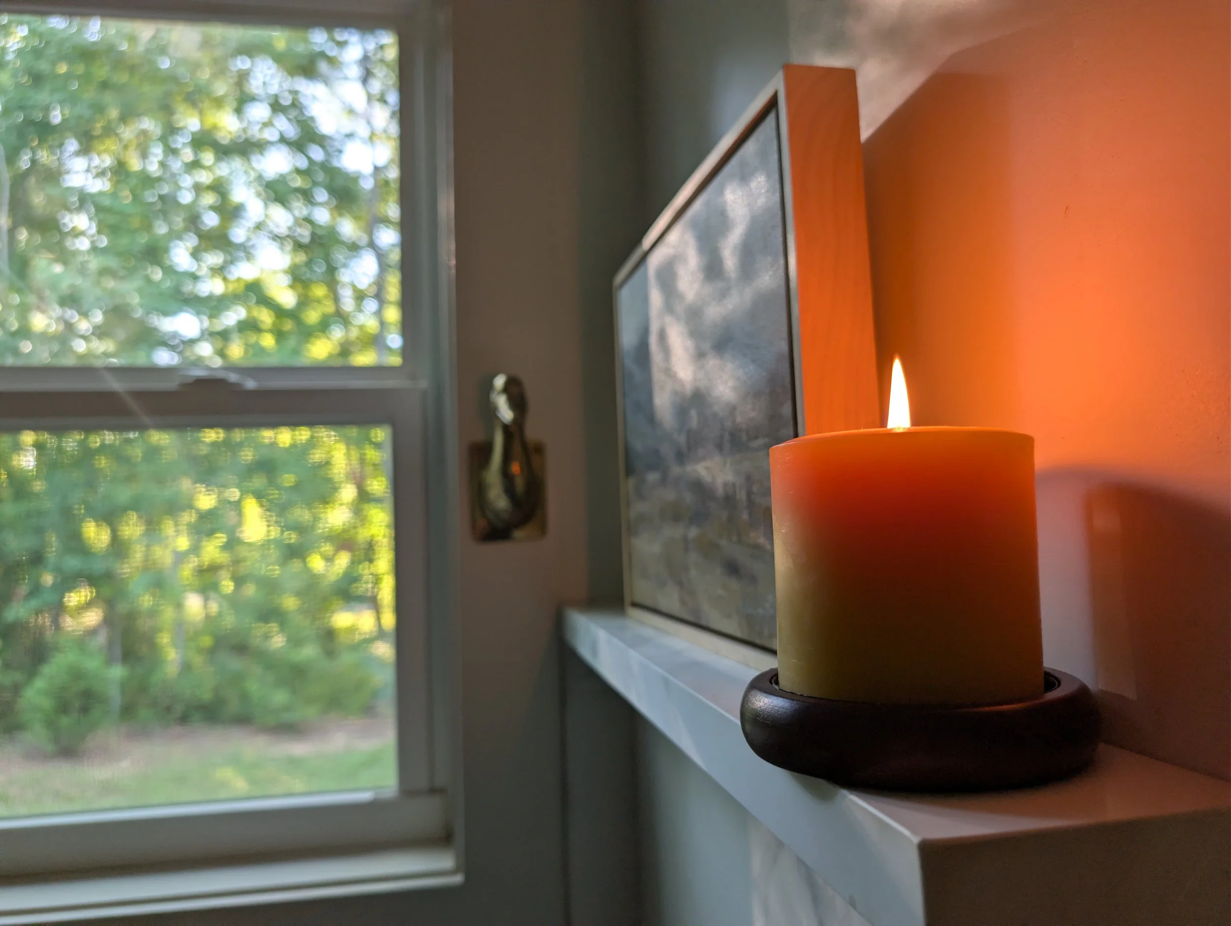 A lit orange candle on a windowsill with a landscape view of green trees outside.