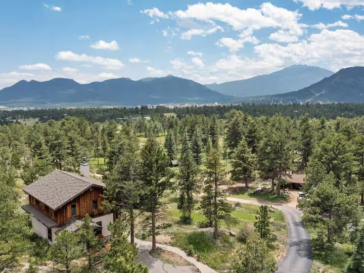 Aerial view of a rural area with a large wooden house surrounded by trees, with mountains in the background and a partly cloudy sky.