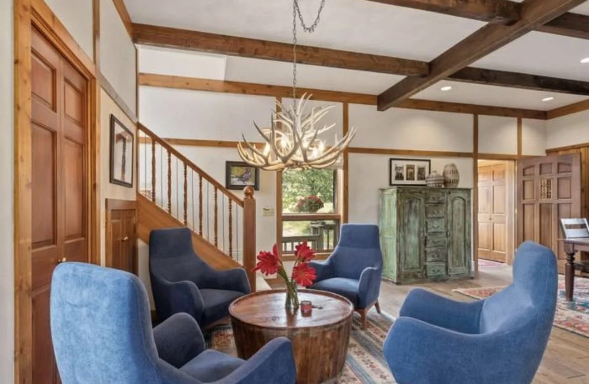 Living room with four blue armchairs around a wooden coffee table, a chandelier made of antlers, a green cabinet, framed pictures, and a staircase with wooden railing.