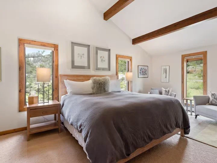 A cozy bedroom with a large bed, gray bedding, and wooden accents. There are windows on either side of the bed, allowing natural light, with framed artwork above the bed and on the walls. Nightstands with lamps, and a small sitting area with chairs a