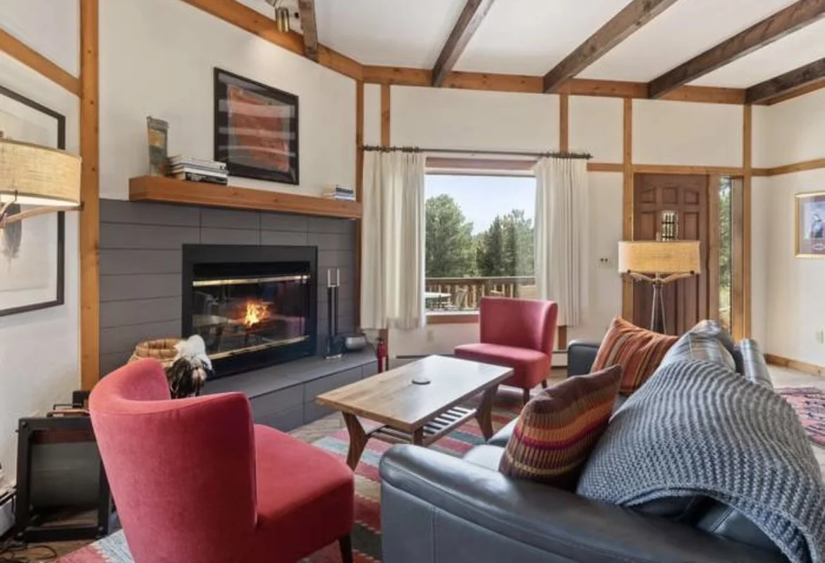 Living room with a fireplace, red armchairs, a grey couch with striped pillows, a wooden coffee table, and a window showing trees outside.