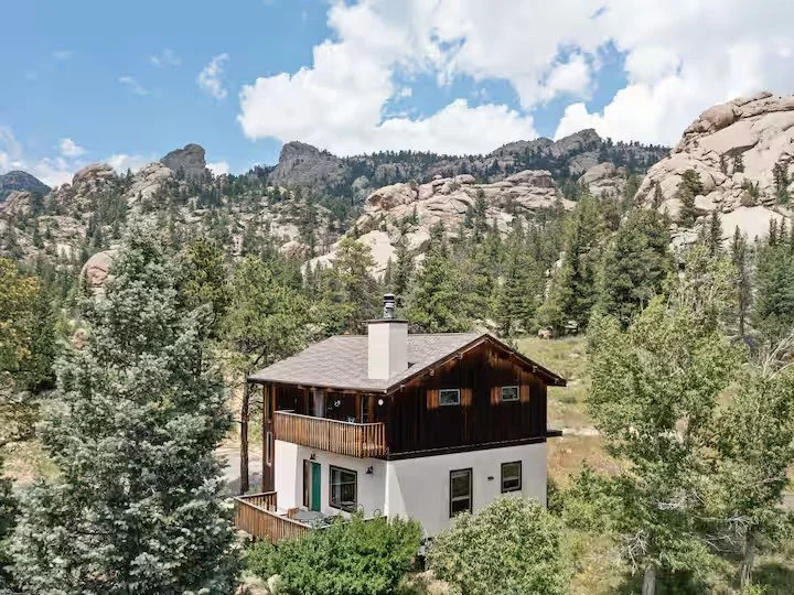 A two-story house with a white lower level and a dark wooden upper level, situated among tall trees with rocky mountains in the background under a partly cloudy sky.