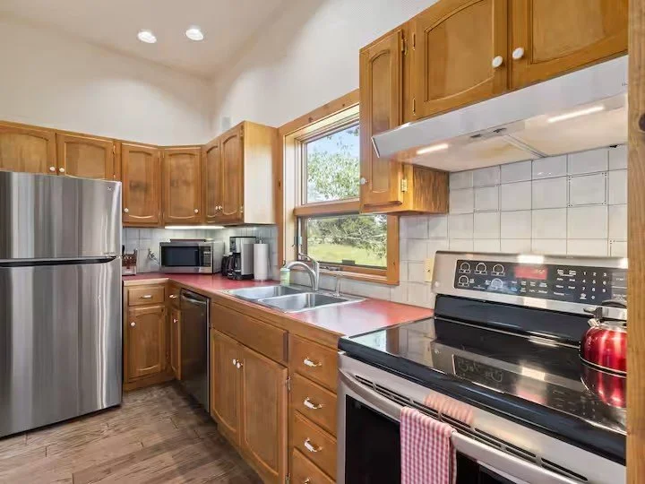 Kitchen with wooden cabinets, stainless steel refrigerator, stove, microwave, toaster, coffee maker, and a red kettle, with a window showing outdoors.