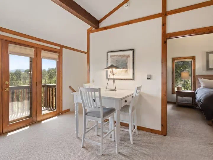 A dining area with a white table and four matching chairs, a framed picture on the wall, and a simple table lamp. To the left, sliding glass doors lead to a balcony with a wooden railing, overlooking trees and a blue sky. Adjacent to the dining area,