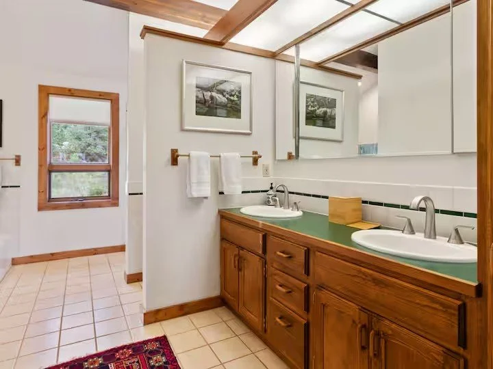Bathroom with a double sink vanity, large mirror, white tile walls, and wooden accents. There is a window with a wooden frame and a partial view of a tiled floor.