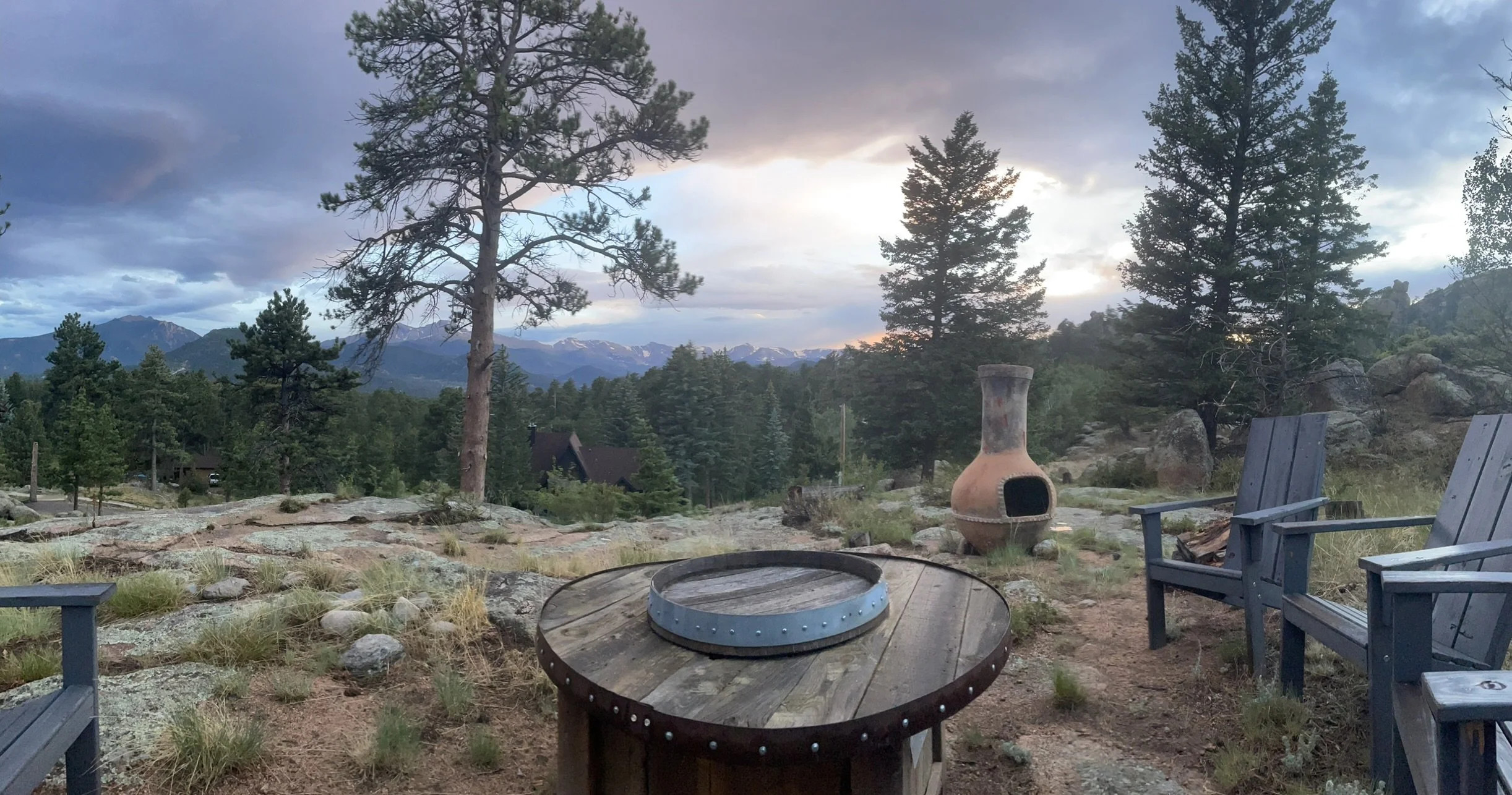 Outdoor patio area with wooden chairs, a round fire pit, and a clay outdoor fireplace, surrounded by trees and mountain views at sunset.