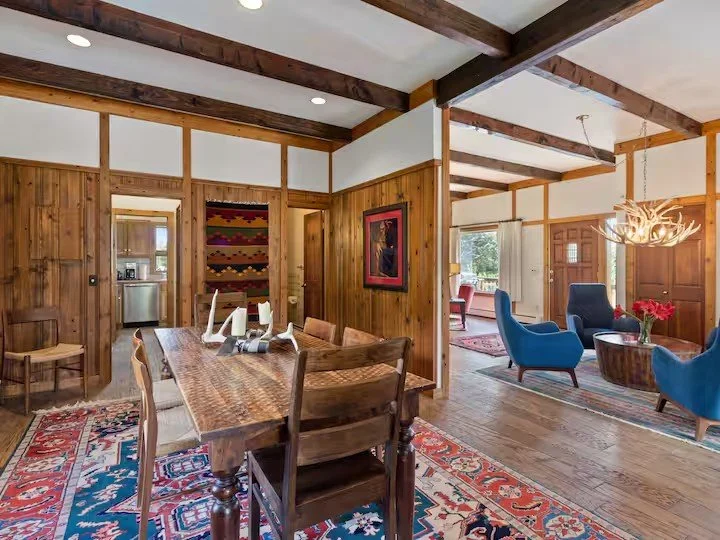 Interior of a rustic living and dining area with wooden wall paneling, a dining table with chairs, a colorful rug, blue armchairs, a chandelier, and a view of a sitting area with large windows.