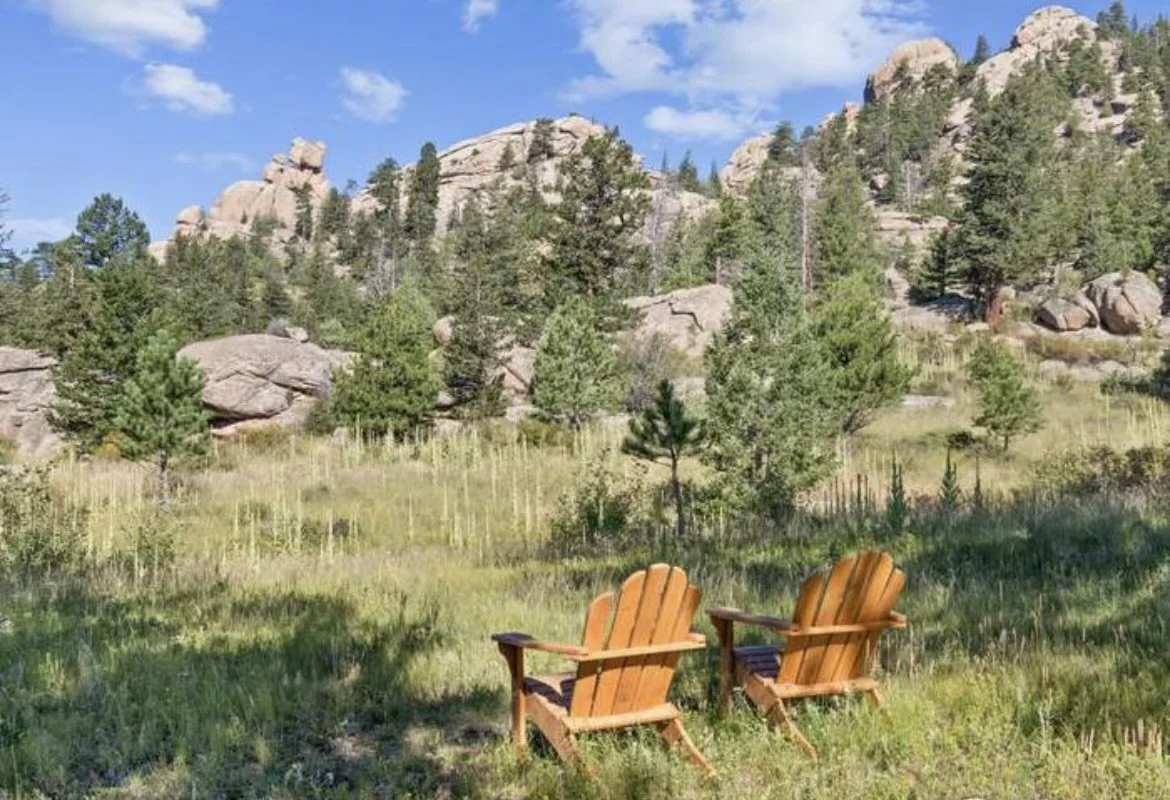 Two wooden Adirondack chairs facing a scenic mountainous landscape with trees, rocks, and a blue sky.