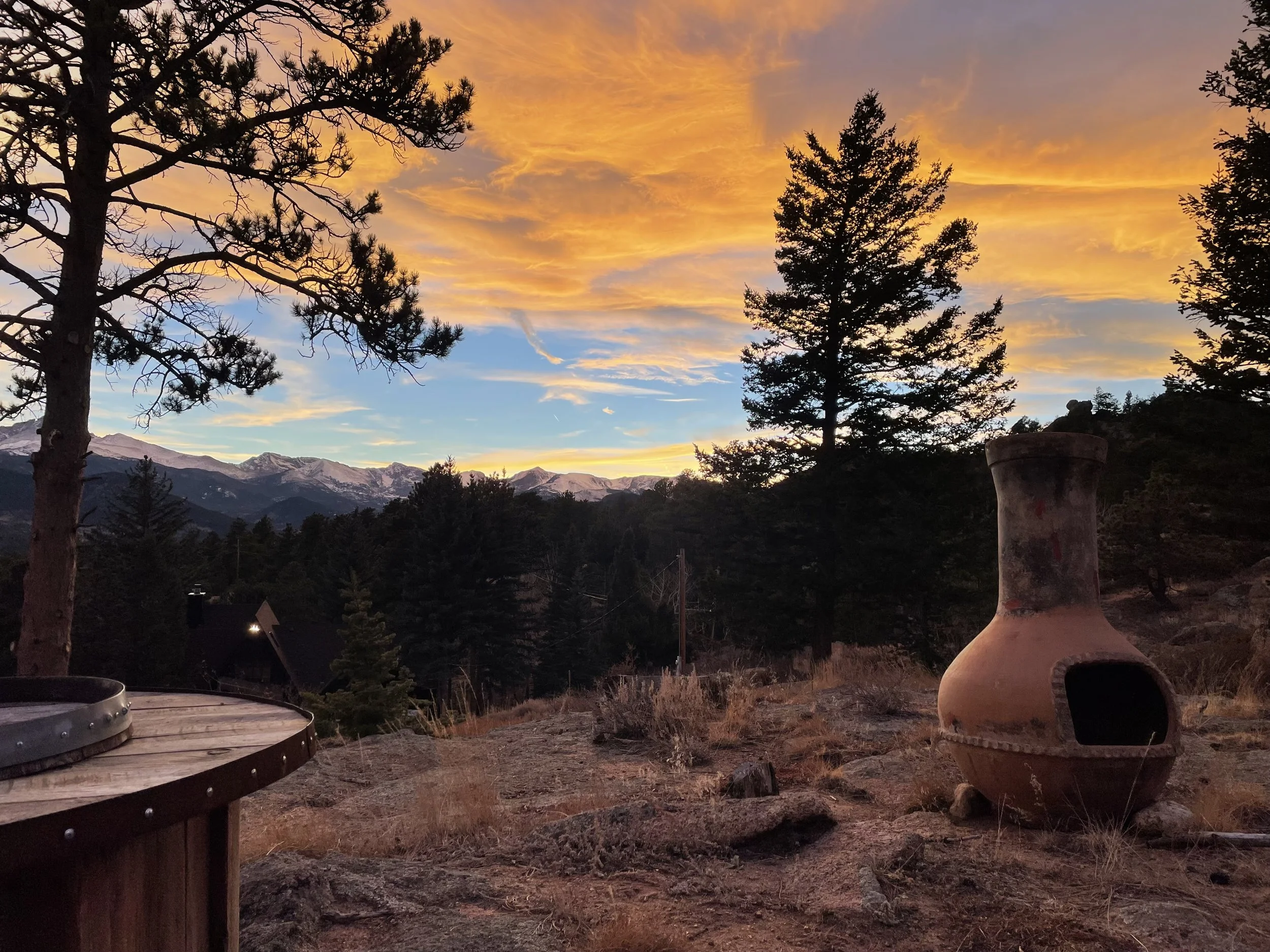 Scenic sunset over mountain range with orange and pink sky, silhouetted pine trees, a large clay outdoor fireplace, and a wooden deck in the foreground.