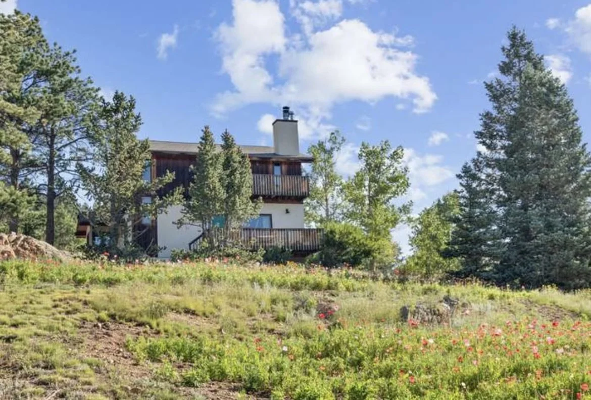 A house on a hill surrounded by trees and a garden with flowers under a partly cloudy sky.