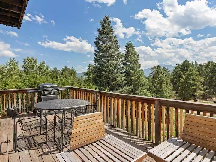 Wooden balcony with outdoor furniture, including a round table with four chairs, two wooden benches, and a grill, overlooking a lush green forest with tall pine trees and a partly cloudy sky.