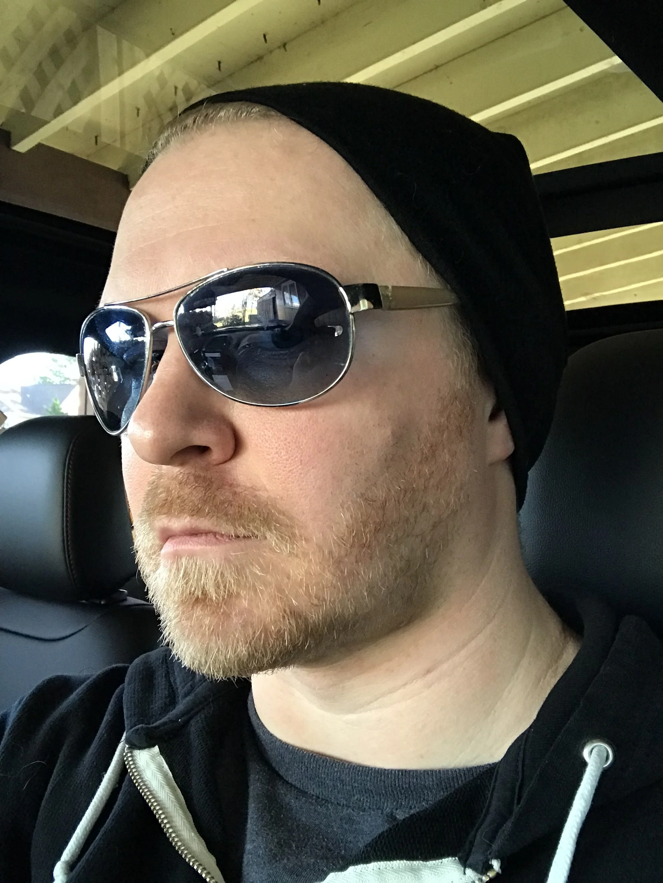 A person with light skin, blonde facial hair, wearing reflective sunglasses, a black beanie, and a black zip-up hoodie with white drawstrings, sitting inside a vehicle.
