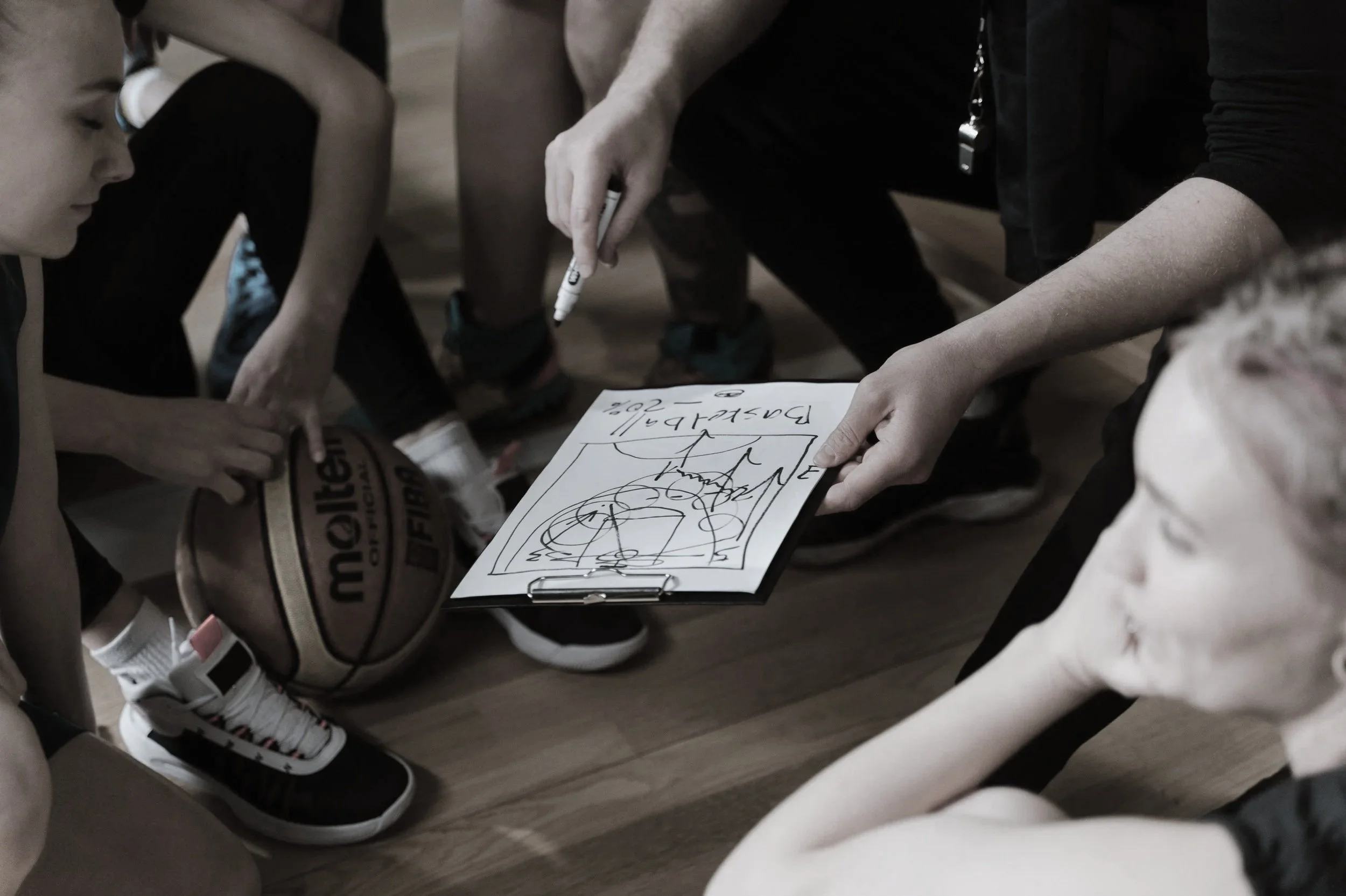 A person is drawing on a clipboard with a marker, showing a basketball and some text, while children in sports jerseys and sneakers watch. The scene appears to be in a gym or sports practice setting.