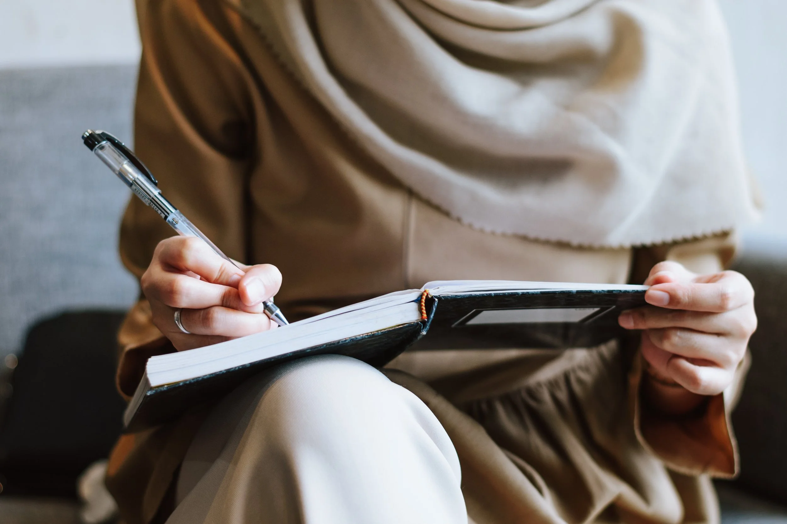 Person sitting down, wearing a beige or tan coat, writing in a small notebook with a black and transparent pen, while holding the notebook with their left hand.