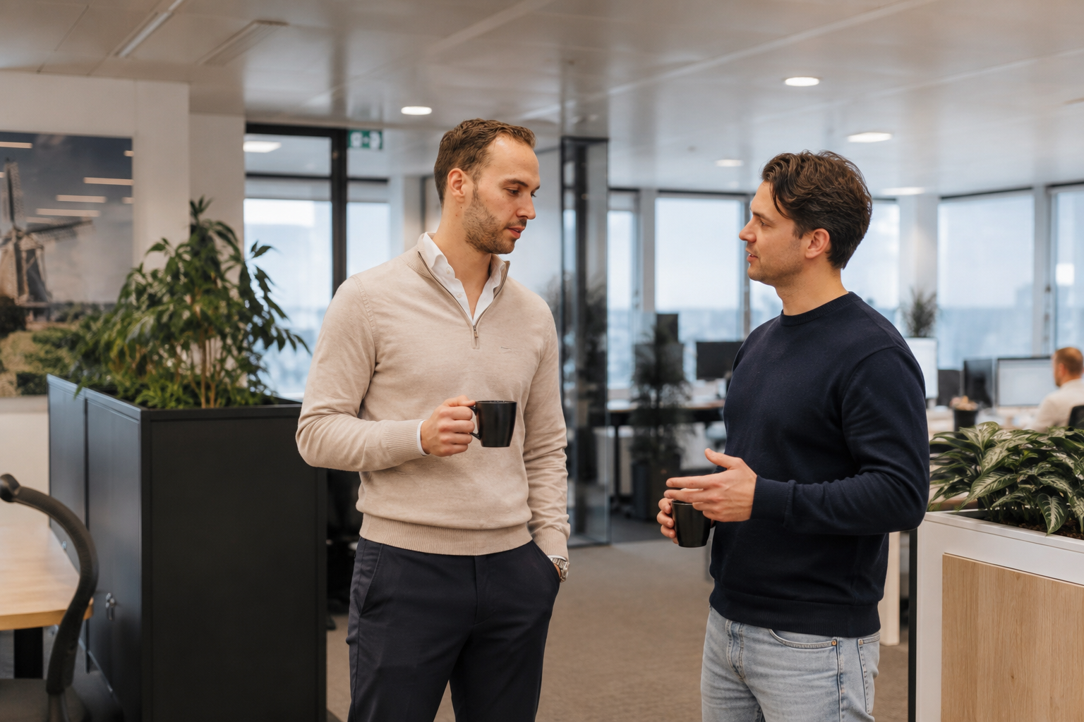 Twee mannen die in een kantoorruimte met planten en bureaus in gesprek zijn, terwijl ze koffie drinken.